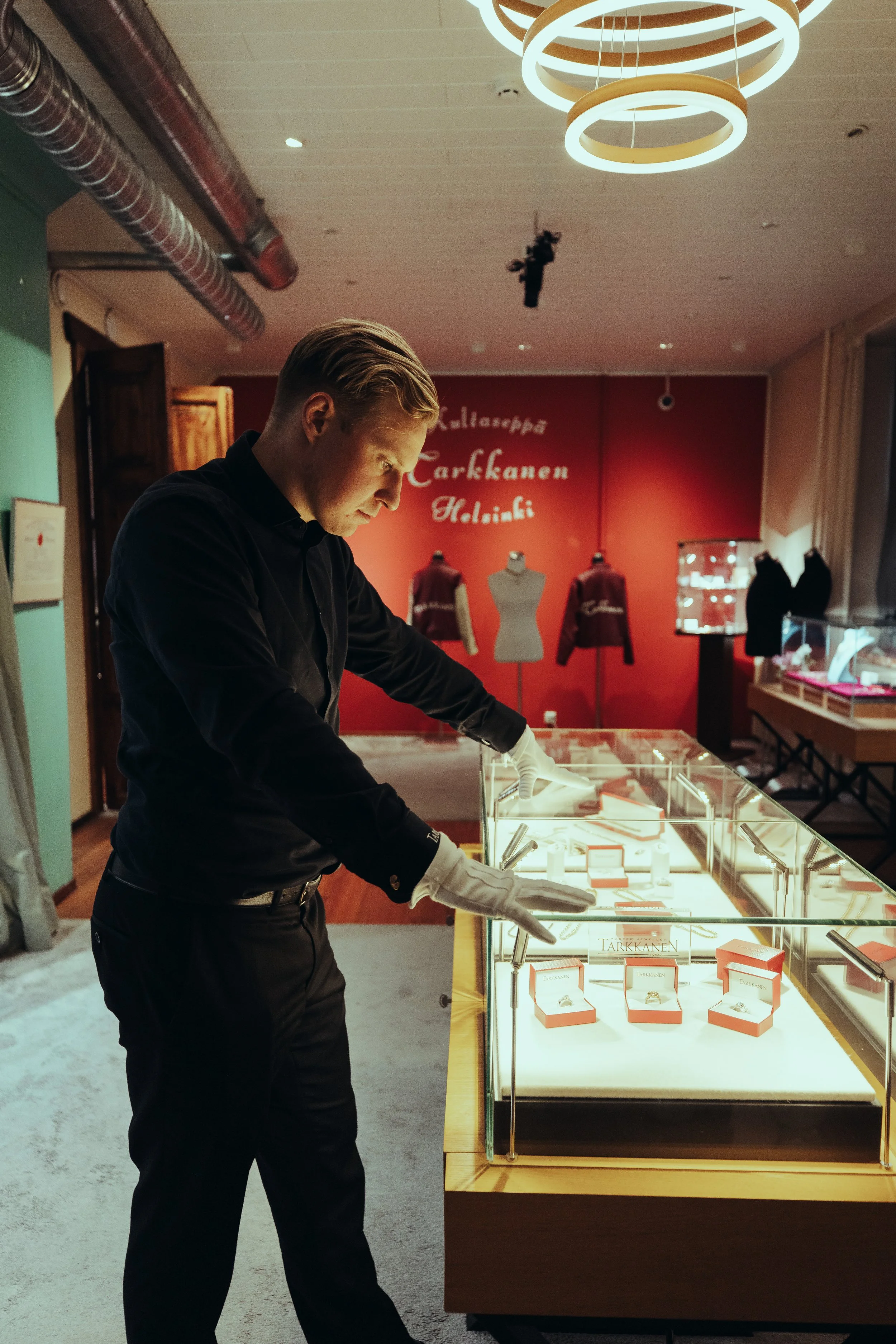 A man wearing black clothing and white gloves looking at jewelry displayed in a glass case at a museum or exhibition.
