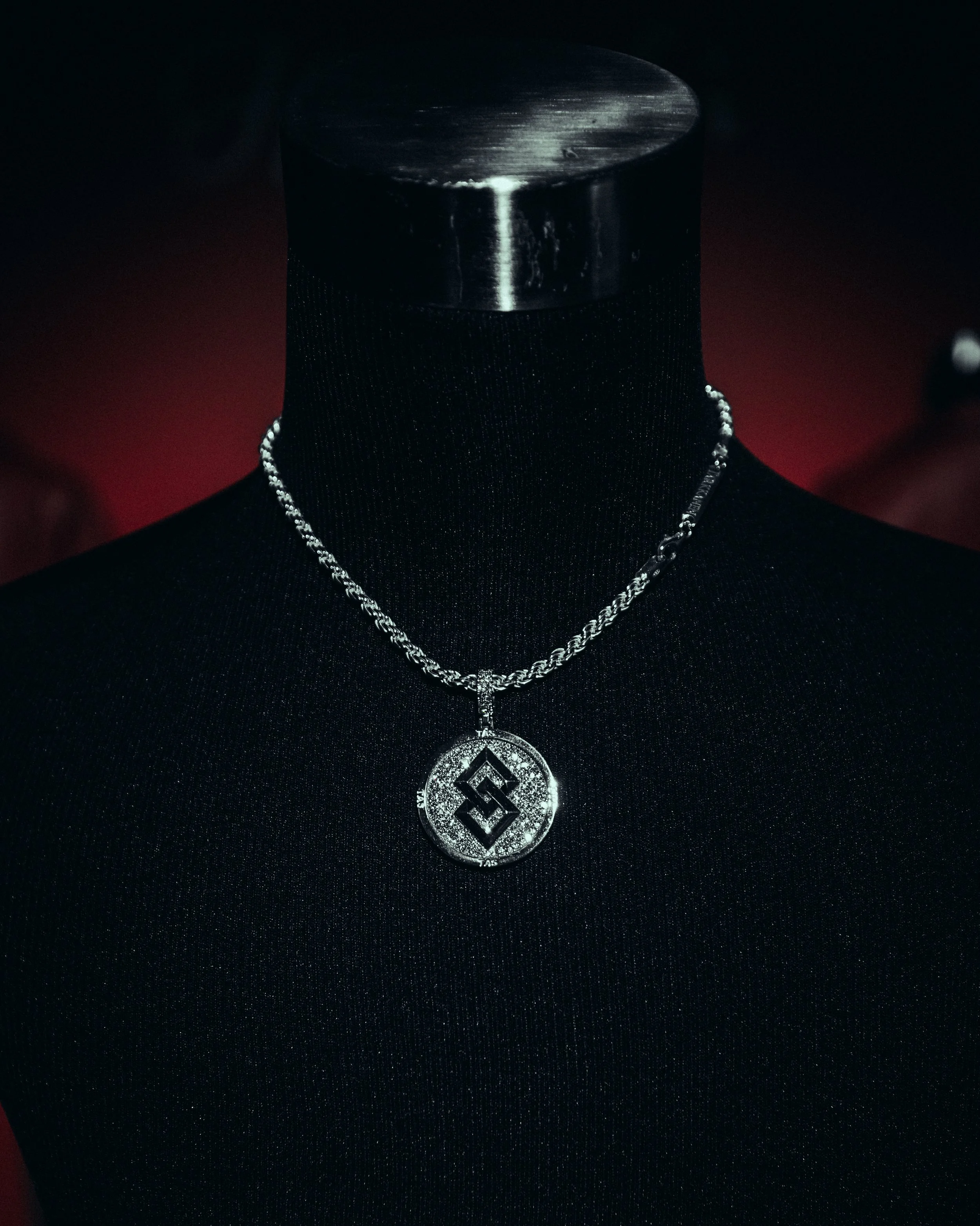 A silver necklace with a round pendant, featuring a geometric design and encrusted with small diamonds, displayed on a black mannequin bust.