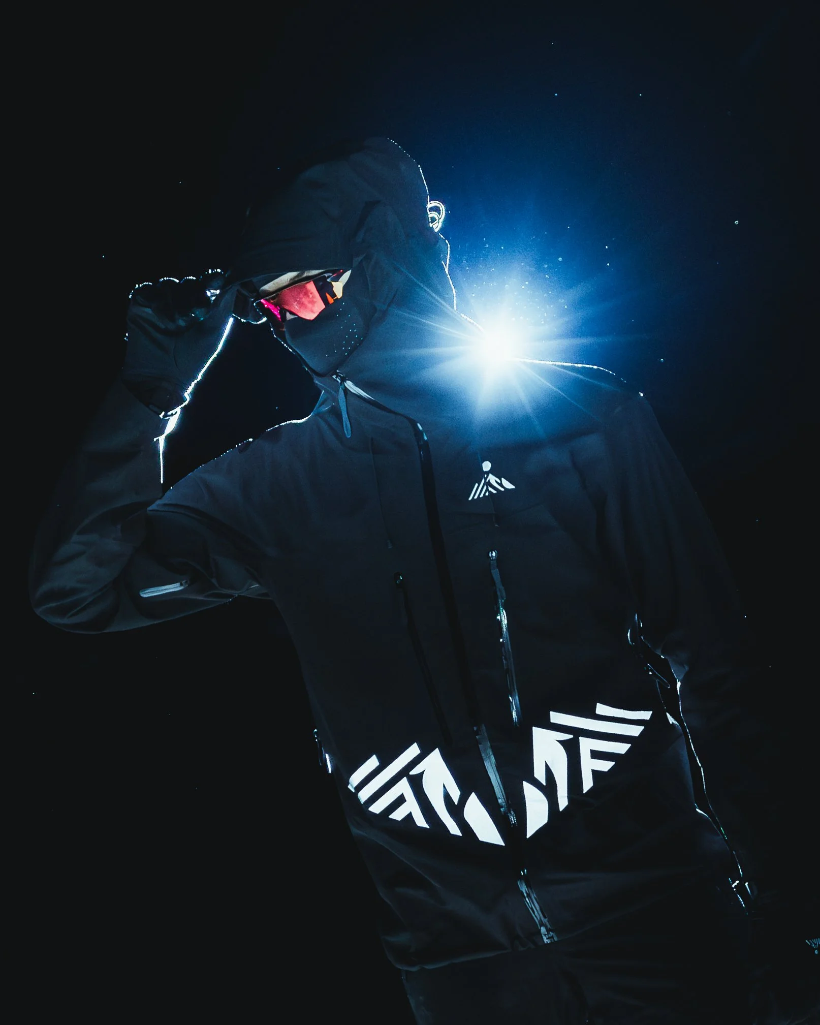 A person wearing black outdoor gear with reflective details, holding a ski pole, outdoors at night with a bright light shining behind them.
