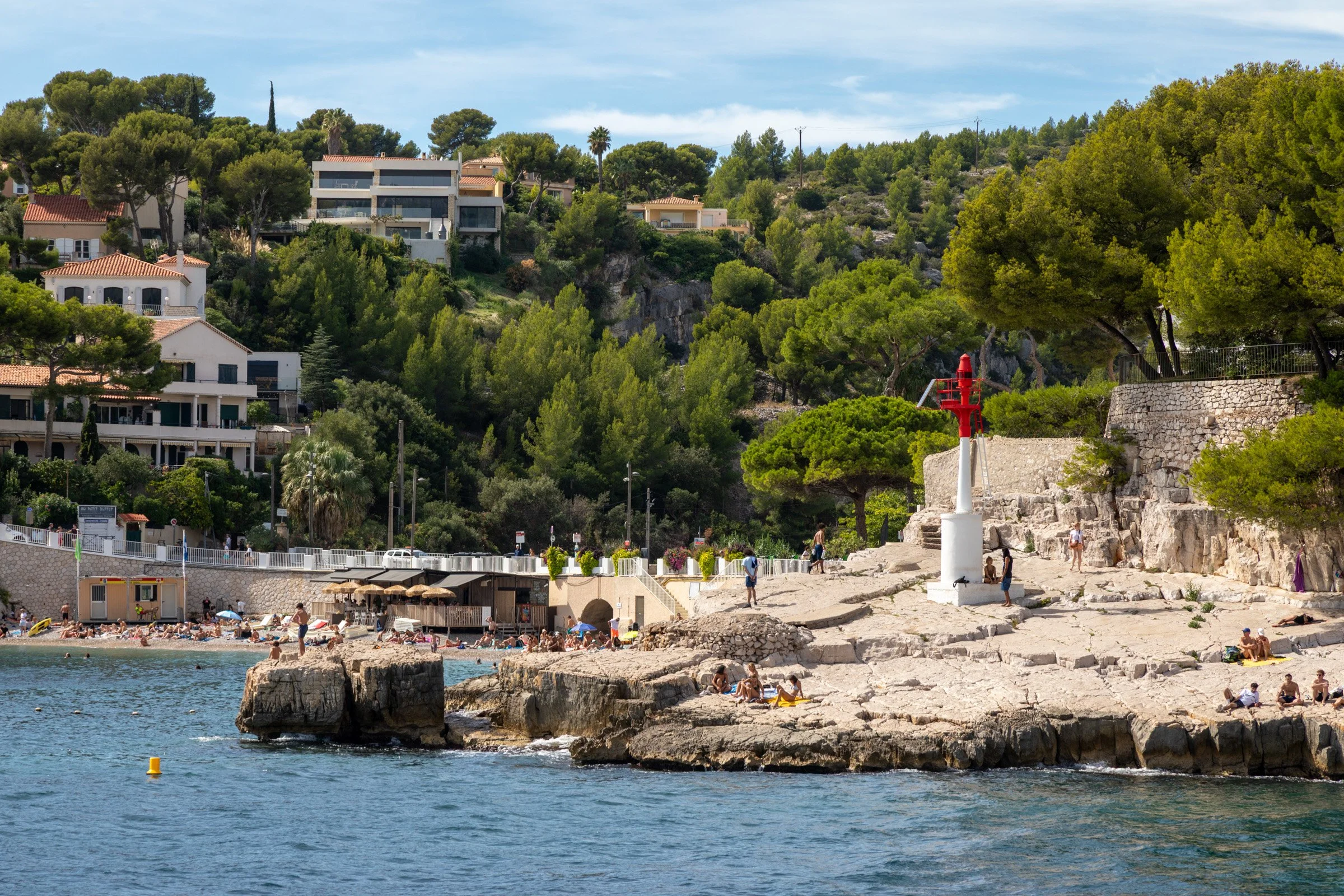 Sunbathers relax on the rocky limestone shoreline of Cassis, France, with a red harbor beacon in the foreground and hilltop villas nestled among Mediterranean pine trees in the background.