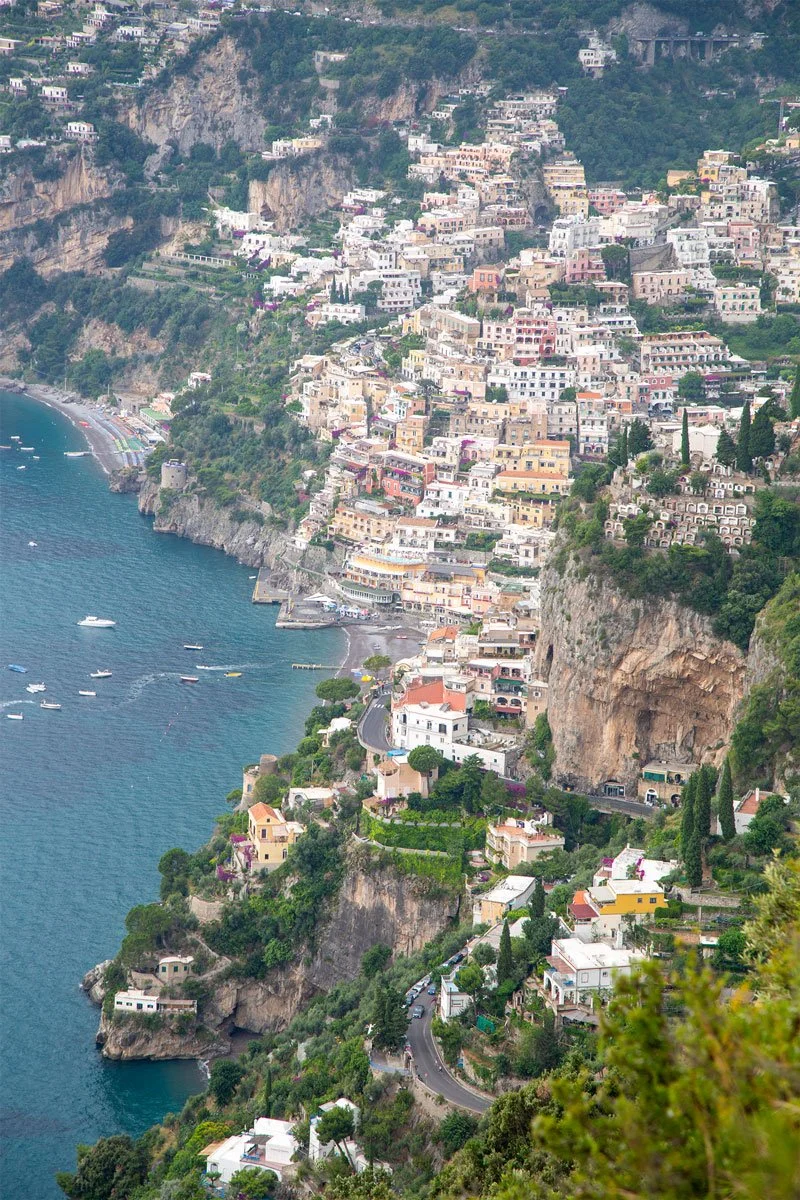 Aerial view of Positano, Italy, a colorful cliffside village cascading down steep rocky slopes to the Tyrrhenian Sea along the Amalfi Coast, with pastel-colored buildings, terraced gardens, and small boats dotting the turquoise water below.