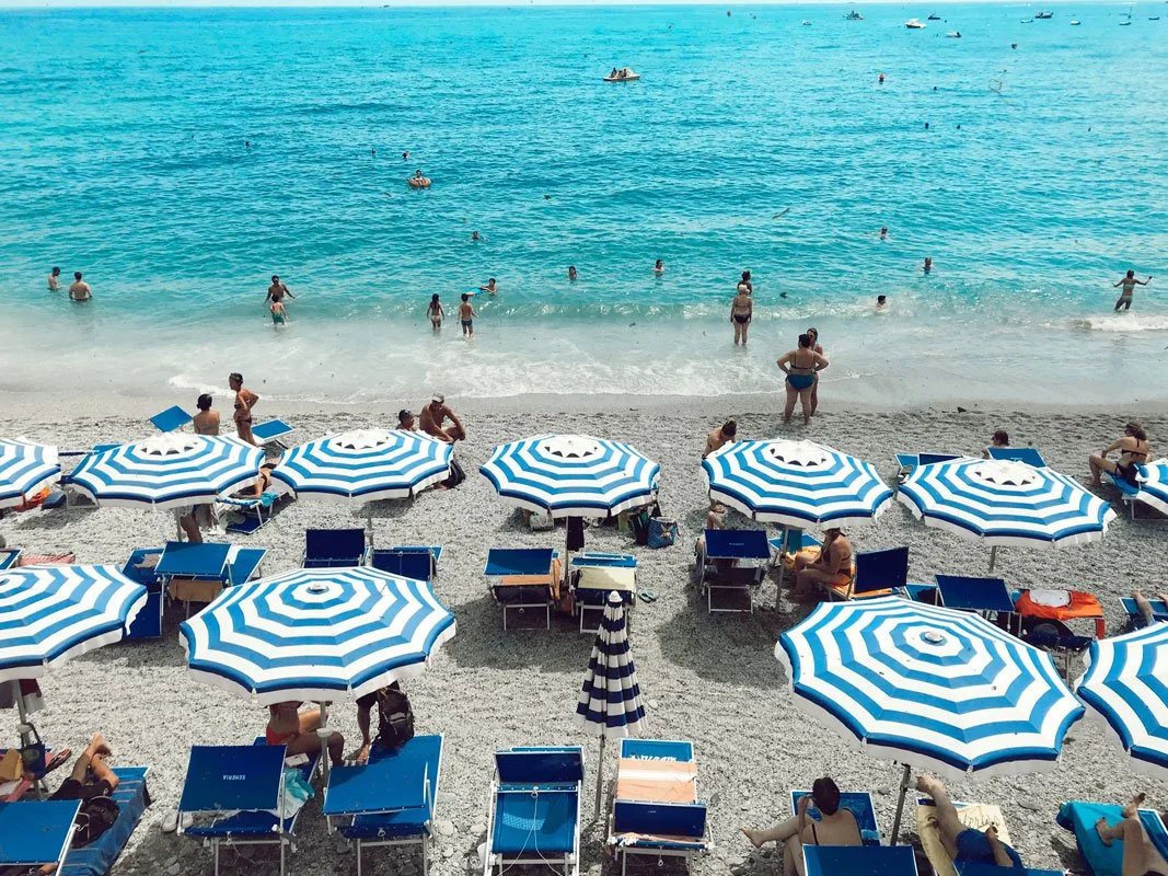 Aerial view of a crowded Italian pebble beach with rows of blue and white striped umbrellas and lounge chairs, as beachgoers relax on the shore and swim in the vivid turquoise Mediterranean Sea.