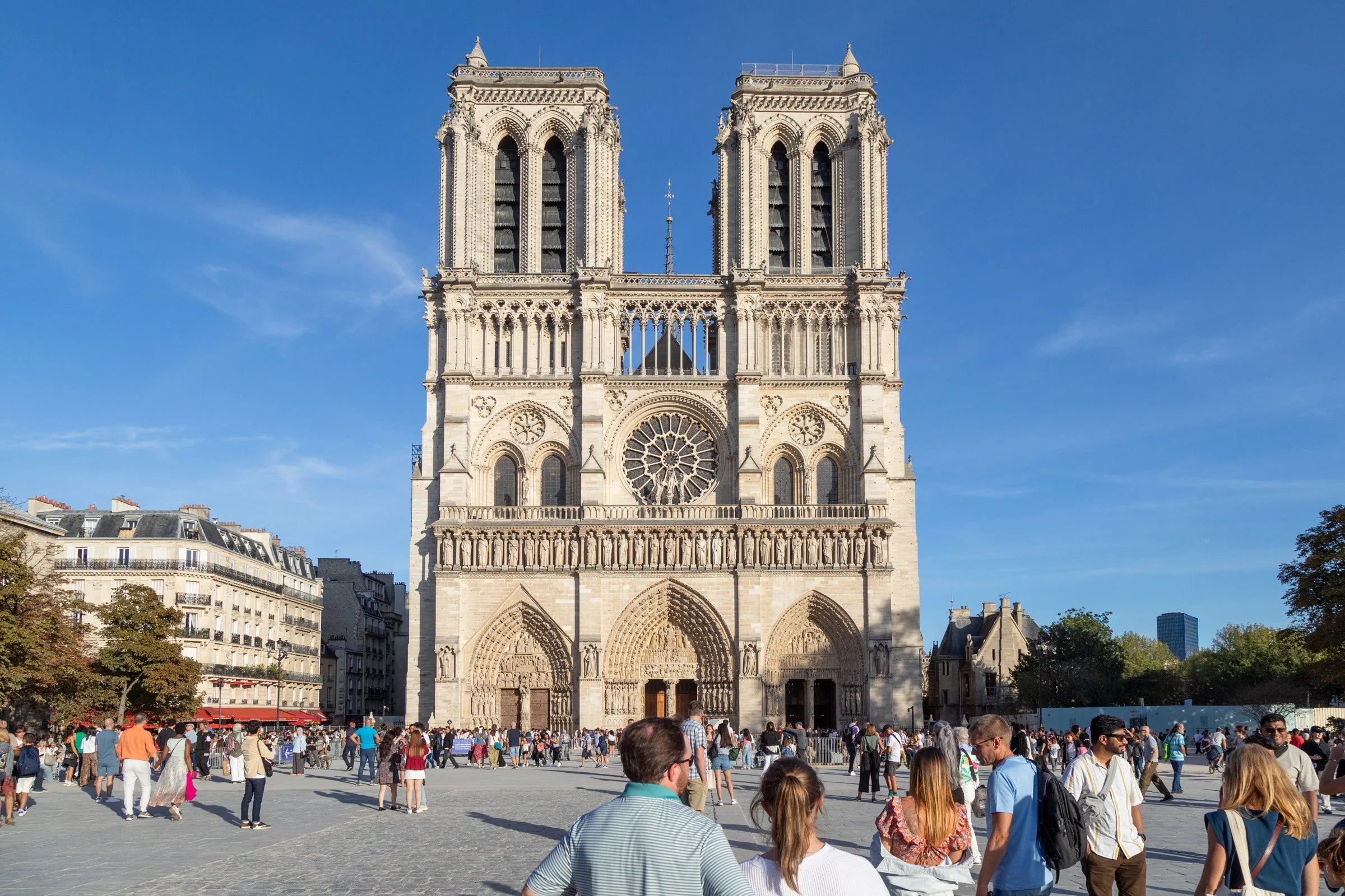 Crowds of tourists gather in the sun-drenched plaza before the iconic Gothic west façade of Notre-Dame de Paris Cathedral, with its twin towers, ornate rose window, and three arched portals set against a vivid blue sky.