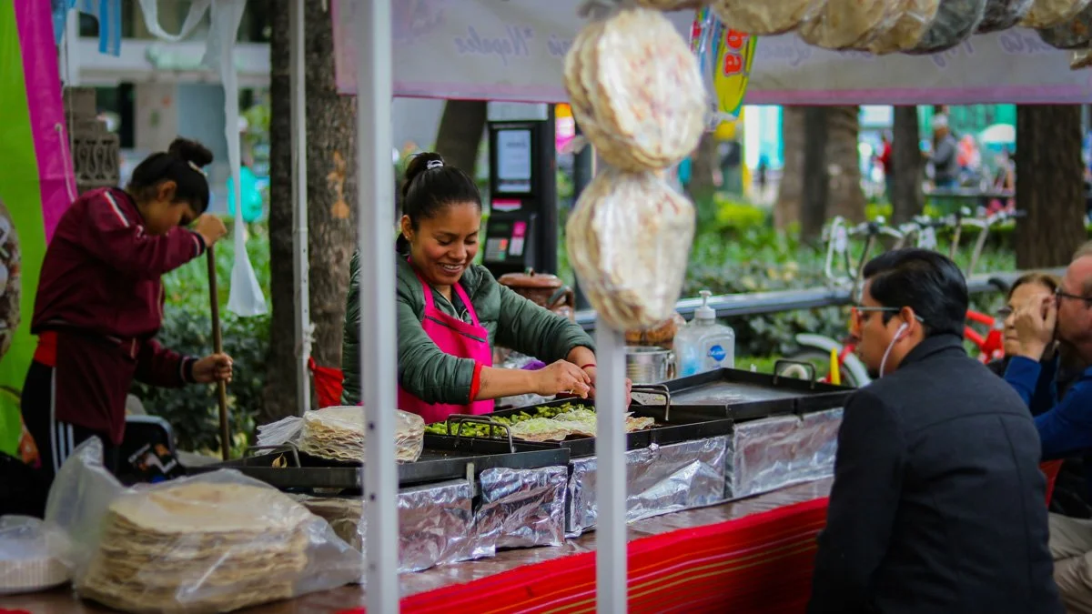 A smiling woman in a pink apron and green jacket prepares artisanal food at an outdoor street market stall, surrounded by tortillas and fresh ingredients. Customers are seated nearby at the colorful red-draped counter.