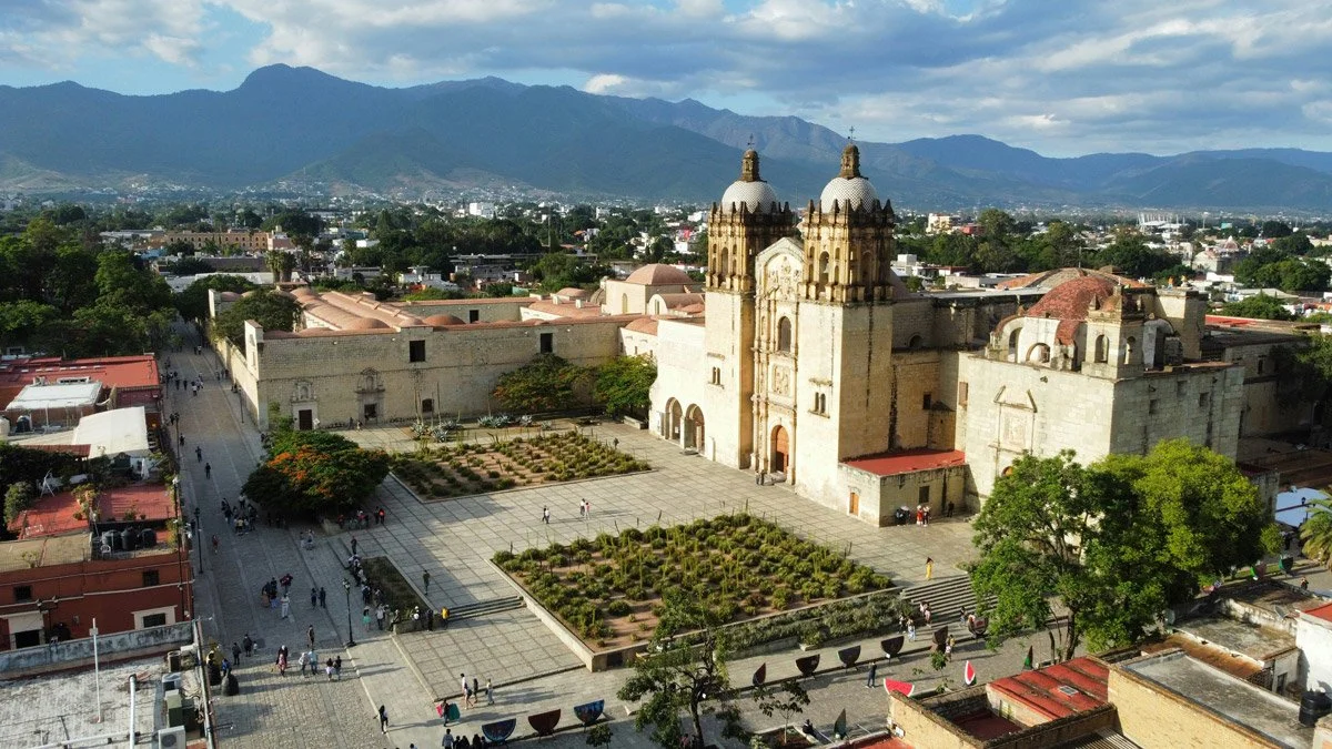 Aerial view of the ornate Baroque facade of the Temple of Santo Domingo de Guzmán and its surrounding plaza in Oaxaca de Juárez, Mexico, with the city and Sierra Madre mountains stretching into the background.