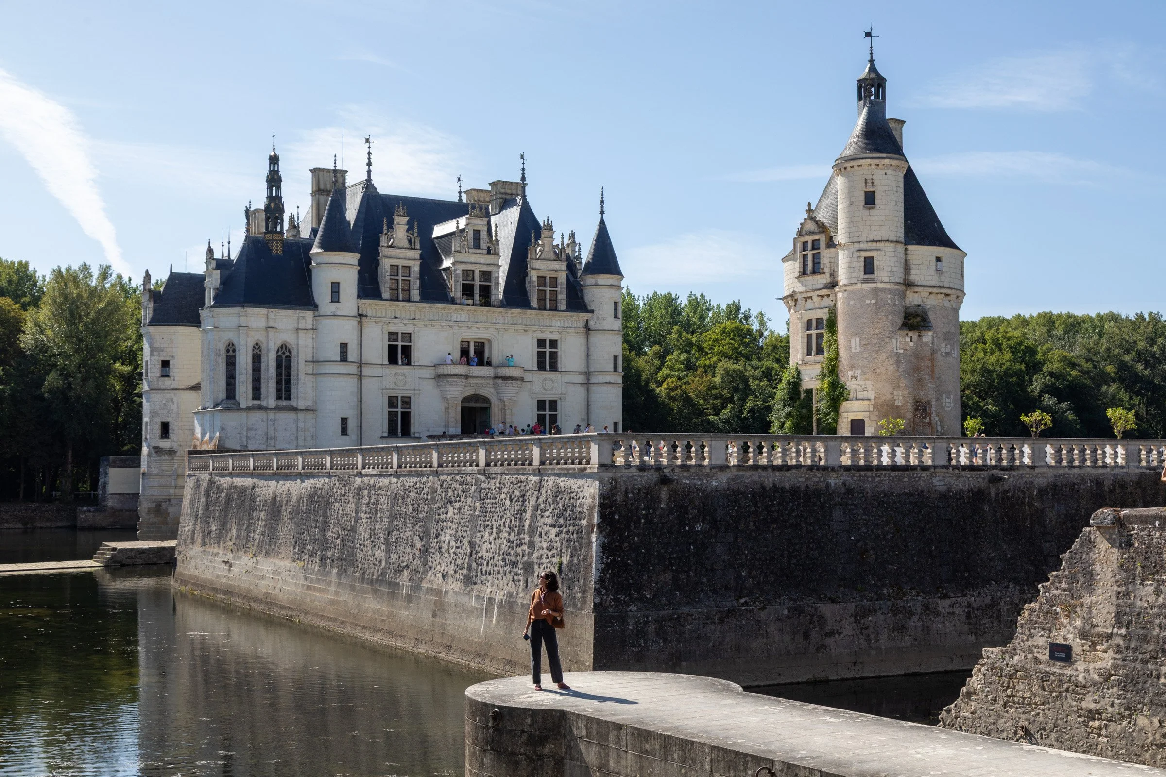 A woman stands on a stone bridge gazing at the iconic Château de Chenonceau, a grand Renaissance castle spanning the Cher River in the Loire Valley, France, on a sunny summer day.