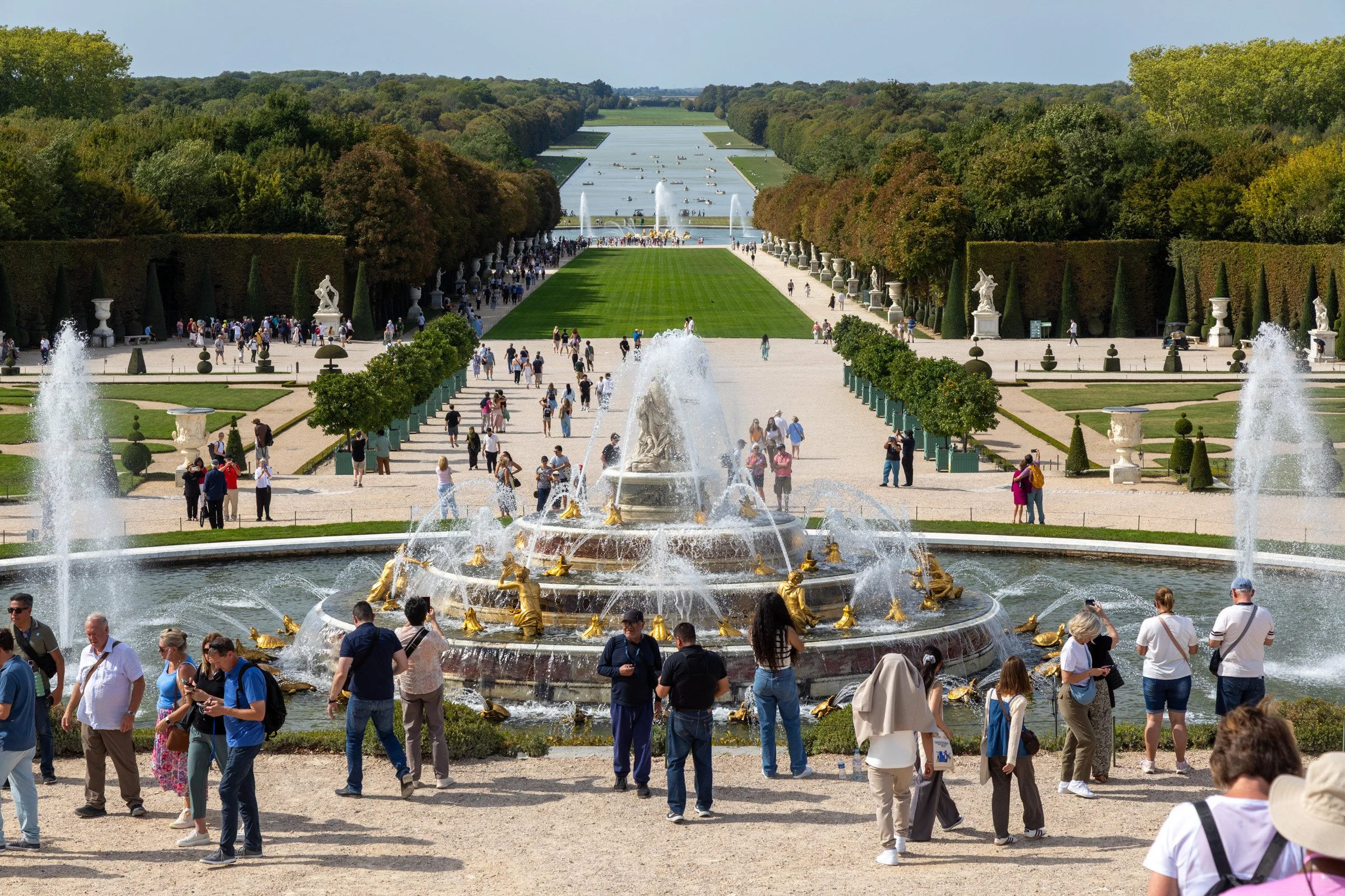 Crowds of tourists gather around the ornate gold-sculpted Latona Fountain at the Palace of Versailles gardens in France, with the Grand Canal and tree-lined promenade stretching into the distance on a sunny day.