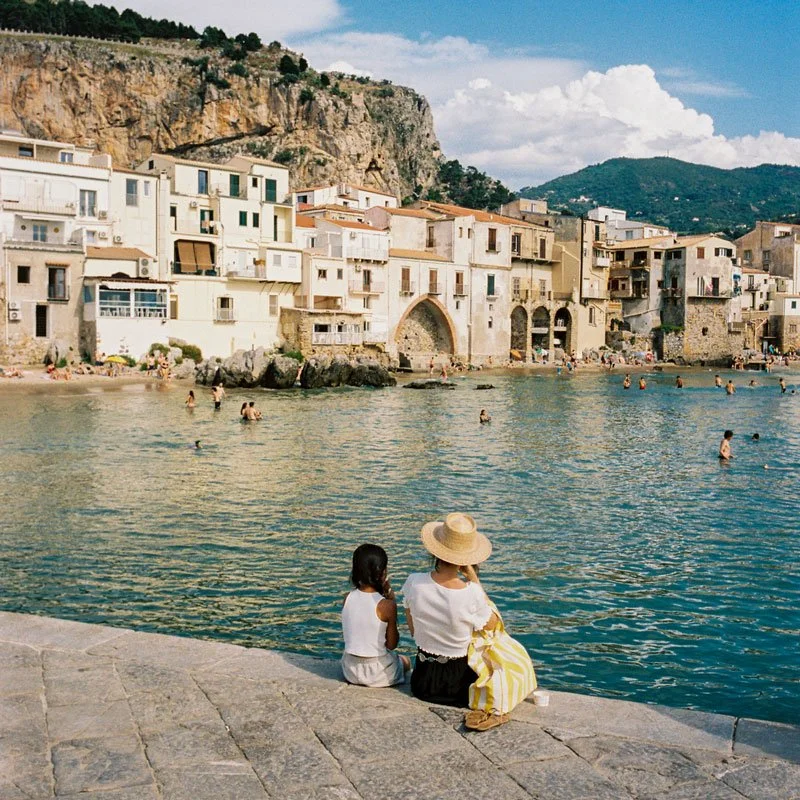 A mother and daughter sit side by side on a stone waterfront promenade in Cefalù, Sicily, gazing out at the turquoise Mediterranean Sea with the town's historic buildings and dramatic rocky cliffs in the background.
