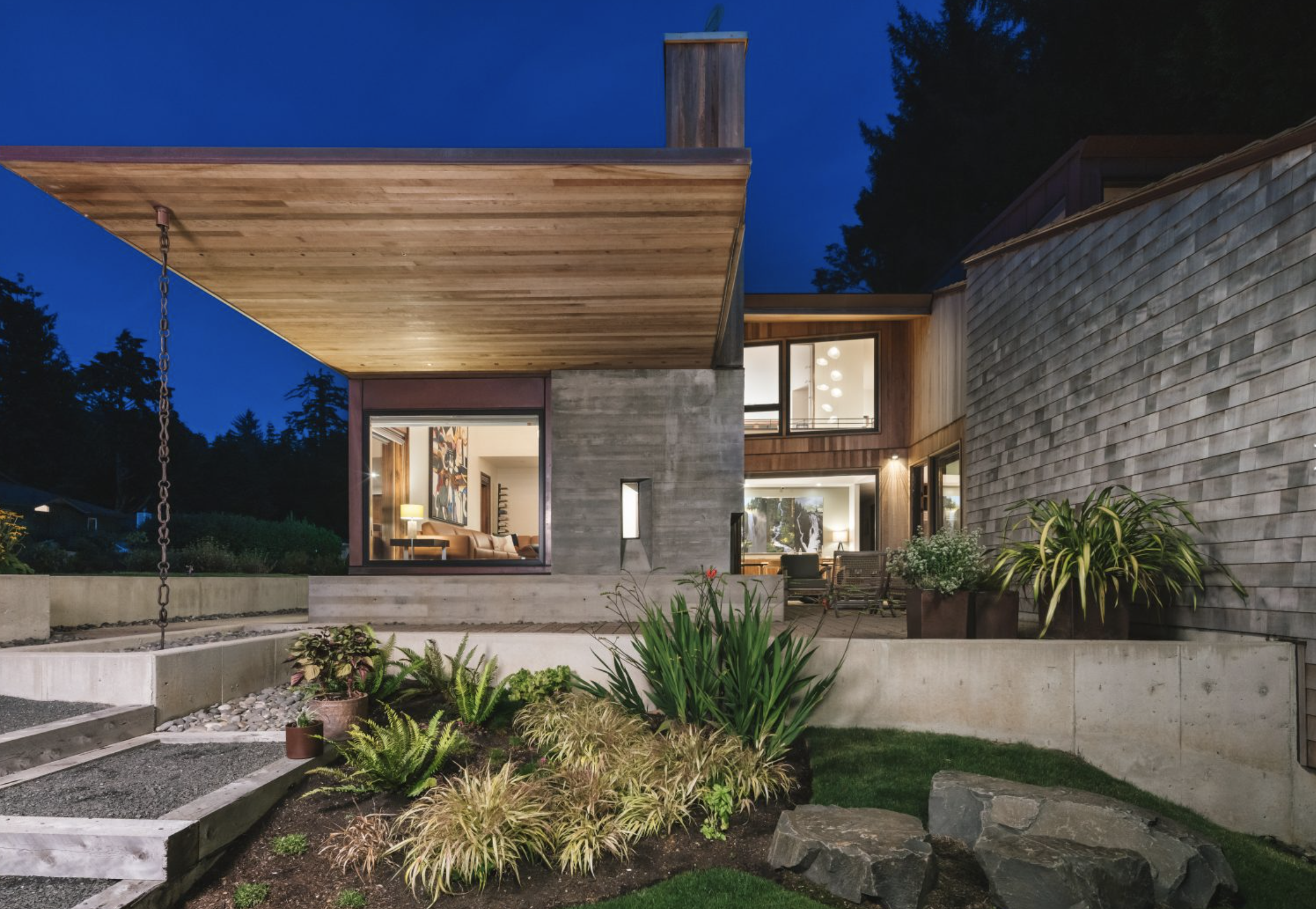 Modern house at night with large windows, outdoor seating area, and landscaped garden with rocks and bushes.