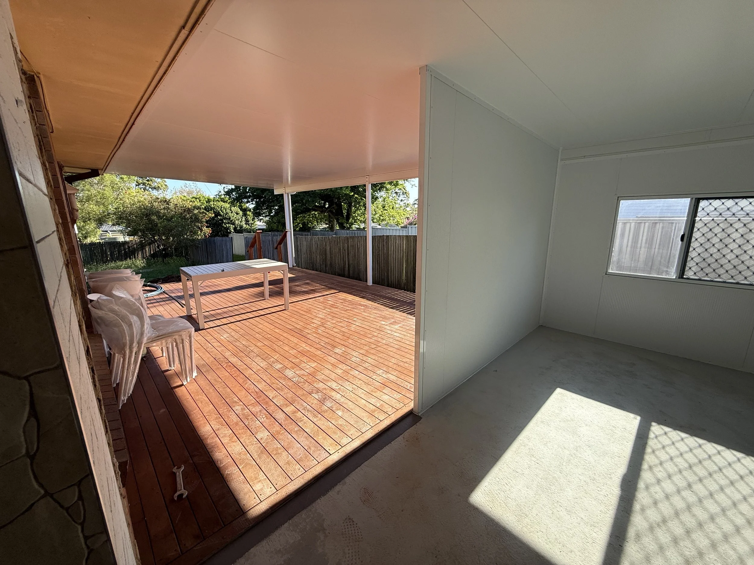 View of a patio with wooden floorboards, white table, and chairs outside, with tree and fence in the background; interior room with white walls and window with security grill, sun casting shadow.