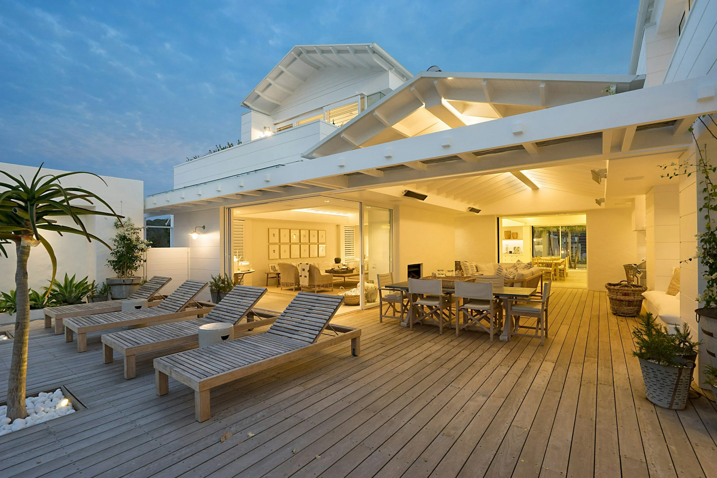 Outdoor rooftop patio of a modern house with wooden decking, sun loungers, potted plants, and a dining area, illuminated warmly in the evening.