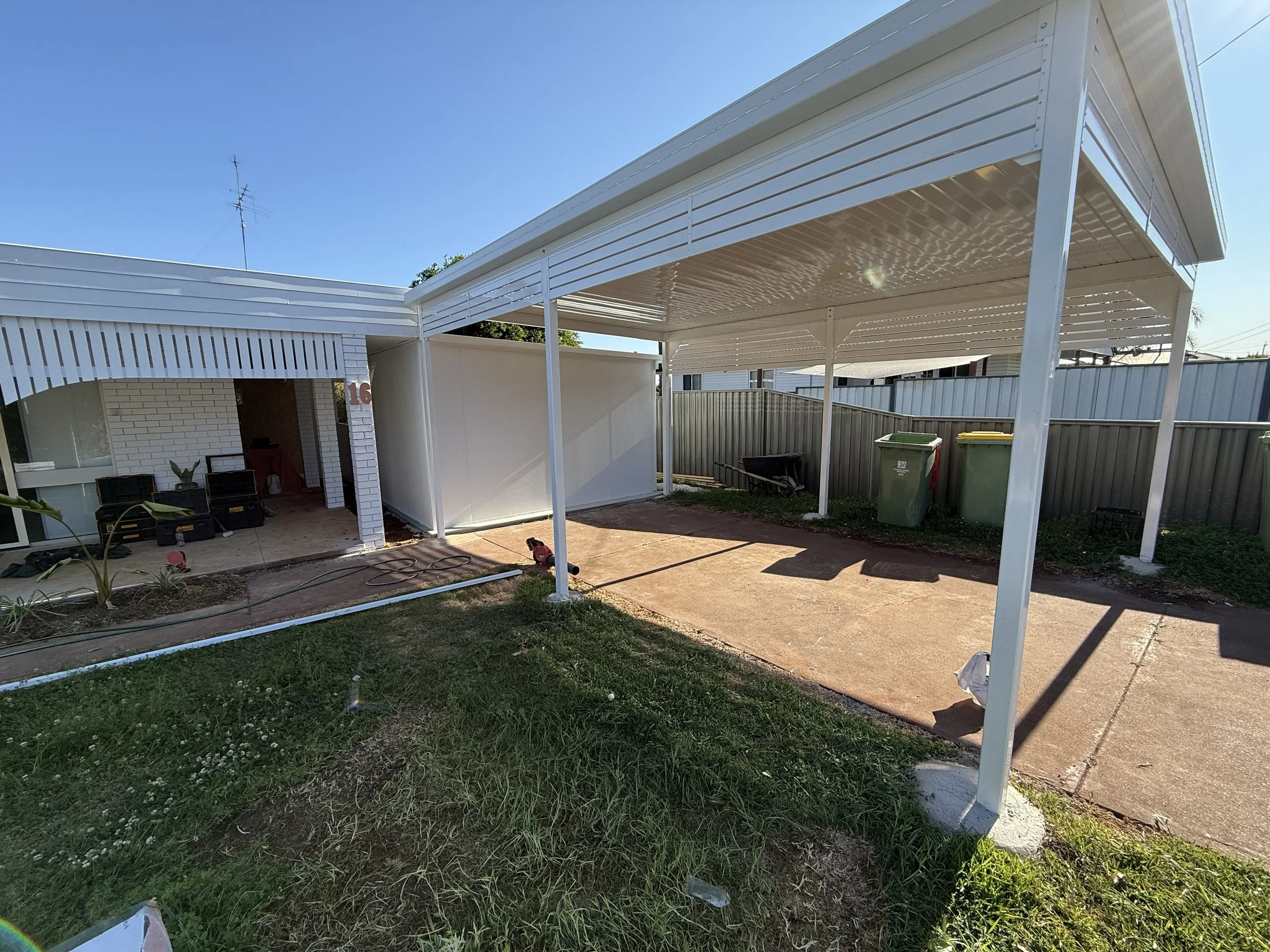 Backyard with a white metal carport, concrete driveway, and fence. There are garbage bins, a wheelbarrow, and some tools and supplies near the house.