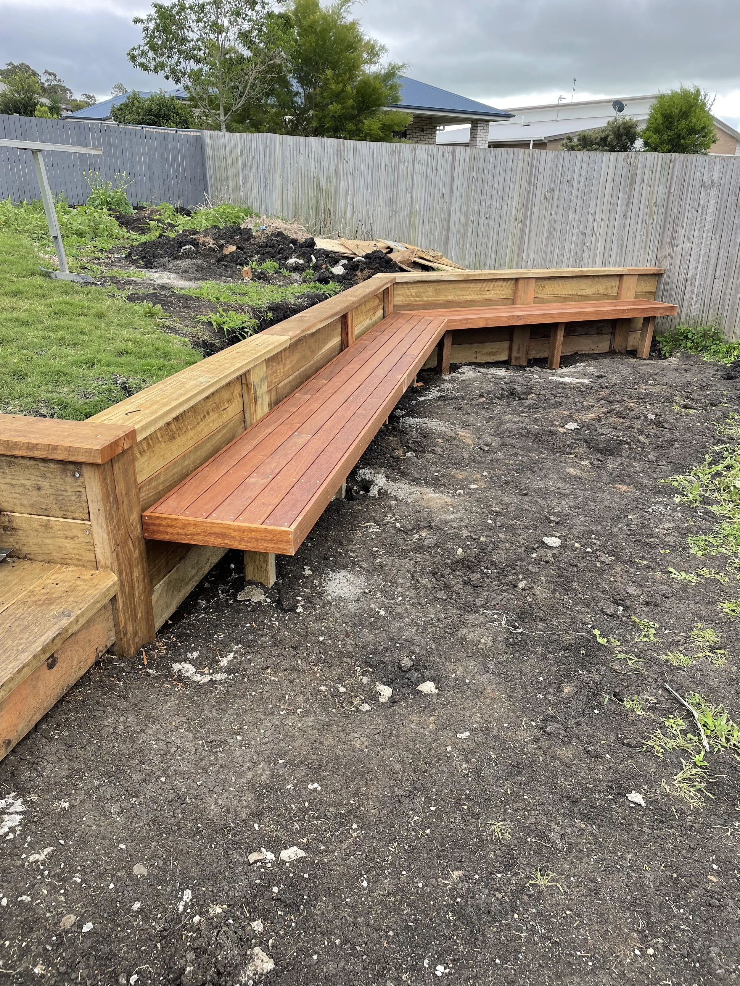 A newly built wooden bench with a backrest in a backyard, with the ground around it prepared for further landscaping or planting.