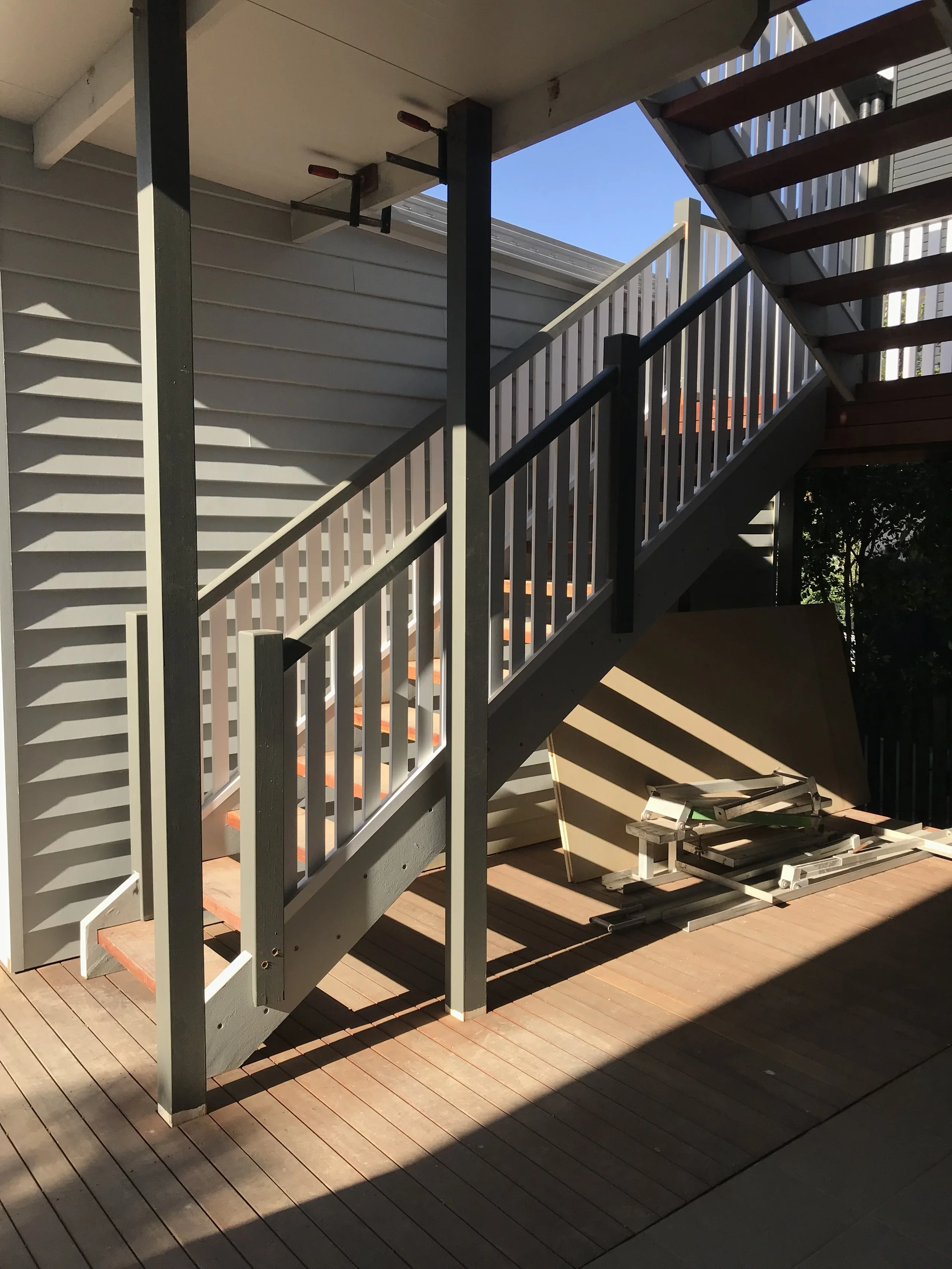 The image shows an outdoor staircase under construction, with wooden steps and a railing, attached to the side of a house with beige siding. Shadows from the staircase and nearby objects are cast on the deck.