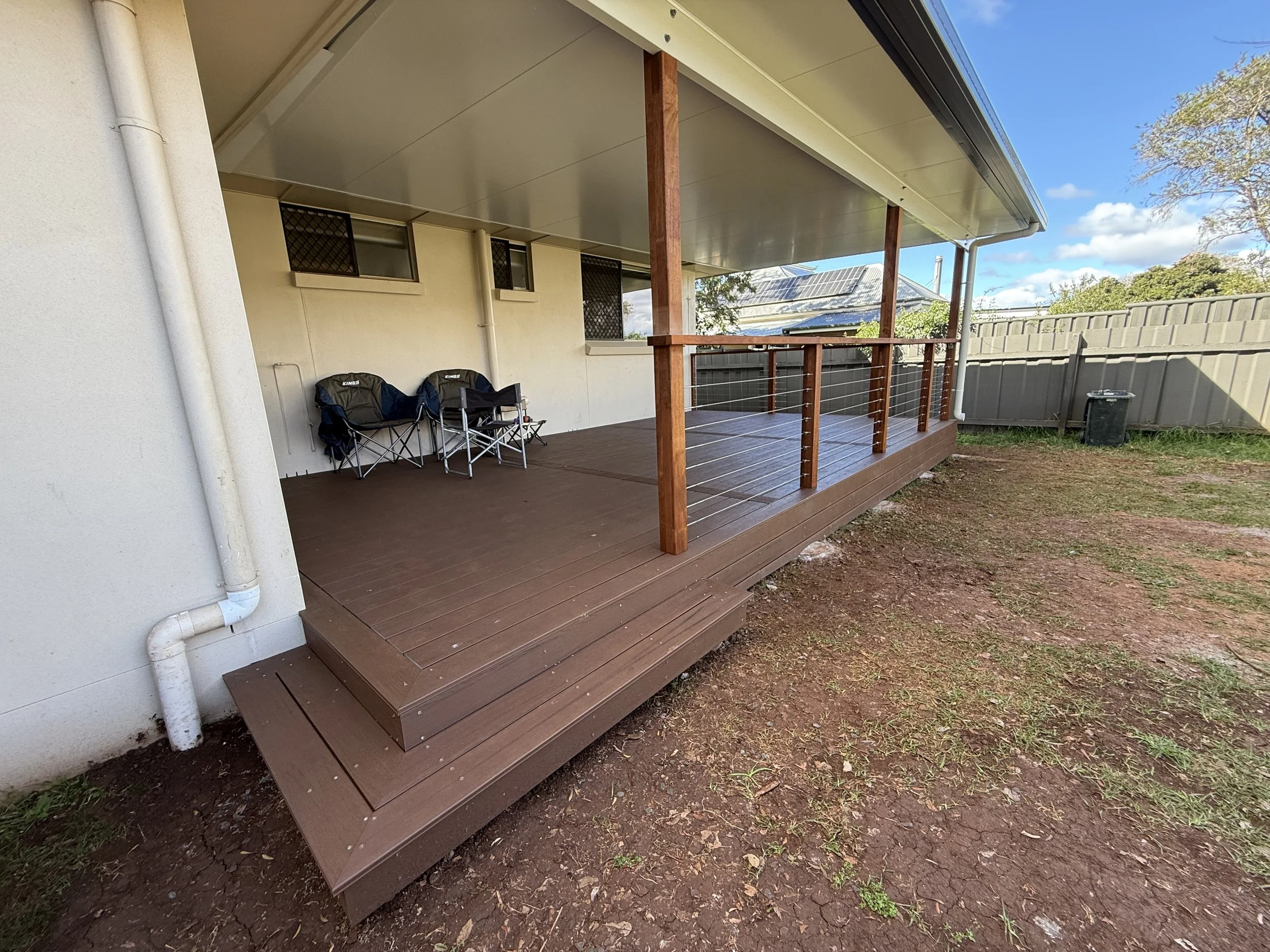 A small covered porch with a brown wooden deck and railing, located at the back of a house. There are two foldable camping chairs on the porch, and a black trash bin in the yard. The yard has patchy grass and dirt, with a gray fence and some trees in the background.