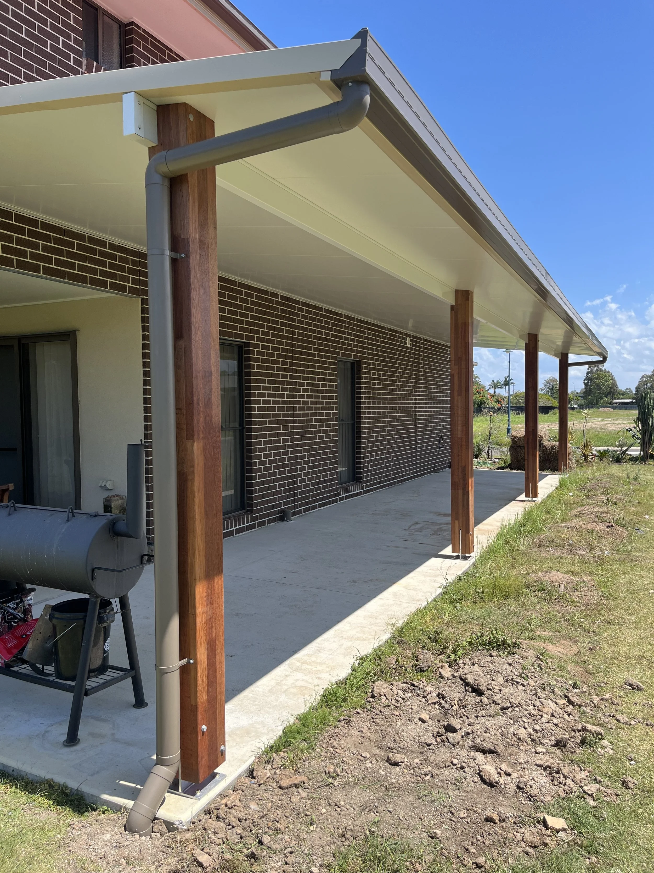 Backyard patio with a concrete floor, supported by wooden and metal posts, under a beige roof extension attached to a brick house with two windows.