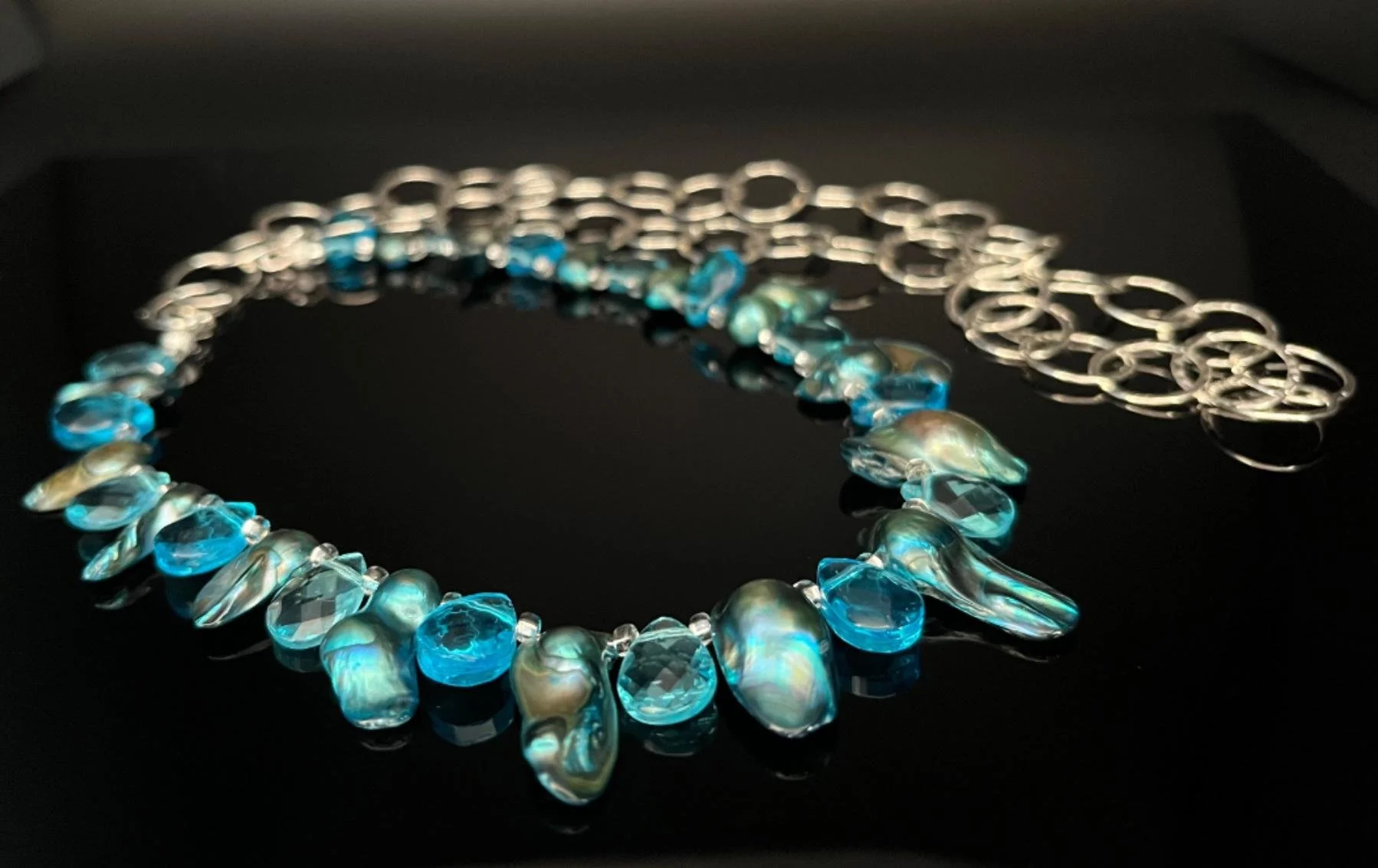 Silver necklace with a chain link design, a heart-shaped pendant, and a teardrop-shaped gemstone at the bottom, displayed against a black background.