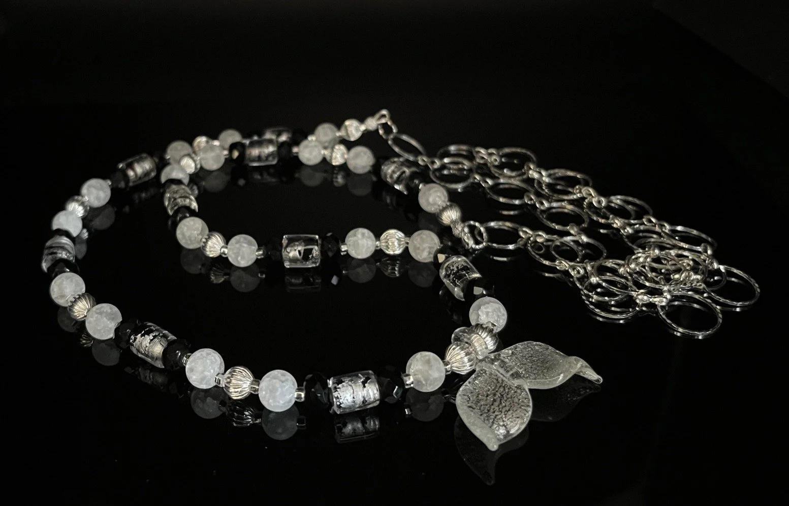 A silver necklace with clear and white beads, including some cube-shaped beads with images inside, displayed on a black reflective surface.