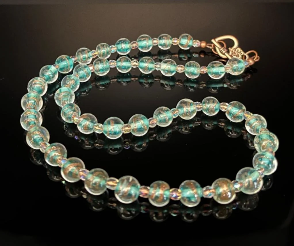 A beaded necklace with green and white marbled beads and white and silver accent beads, resting on a reflective black surface.