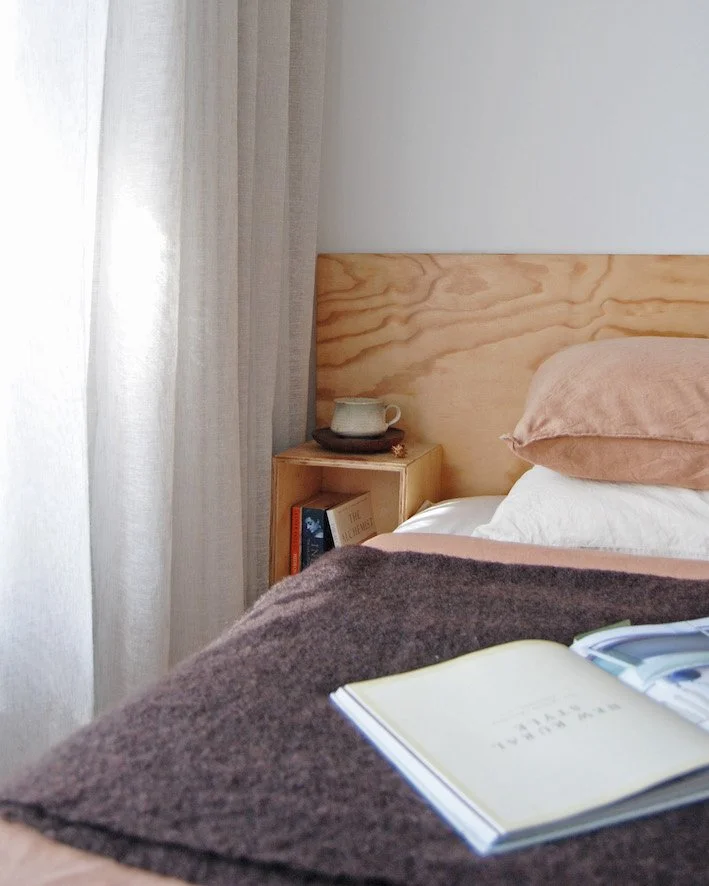 Plywood headboard with linen bedding in dusty pink, charcoal wool throw and a timber side table with ceramic mug — a quiet bedroom corner in a residential home in Newcastle.
