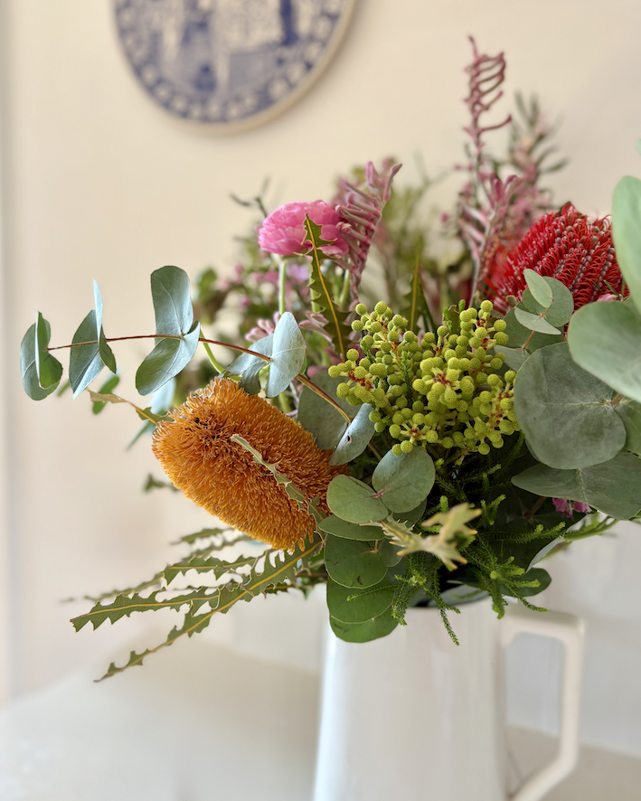 Australian native flowers — banksia, kangaroo paw, eucalyptus and dahlia — arranged in a white jug beneath a blue and white delftware plate. From Olivia Munroe's home in Newcastle.