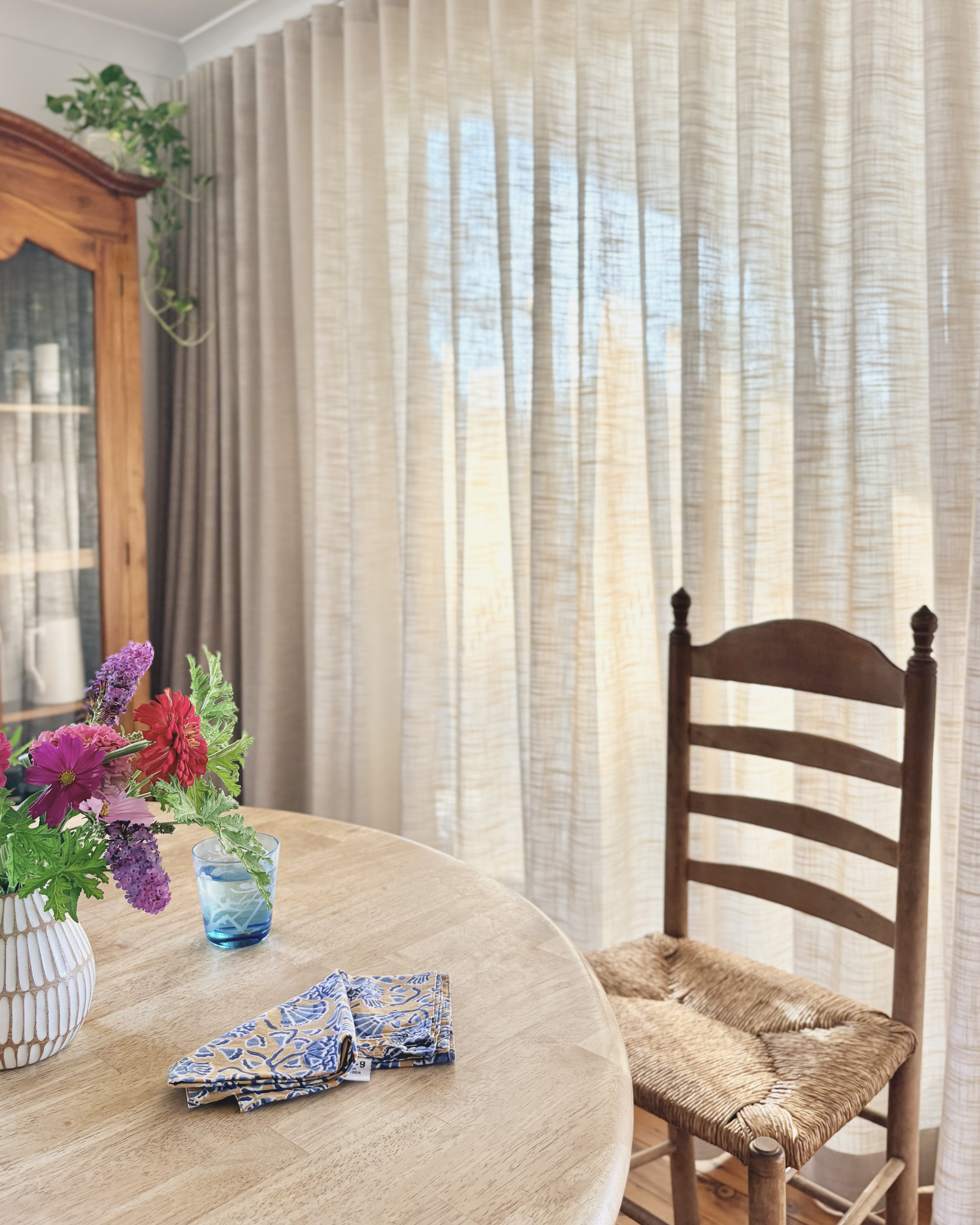 Round timber dining table with antique ladder-back rush chair, sheer linen curtains and Australian native flowers in a white jug. From the Newcastle home of Oliv Interiors.