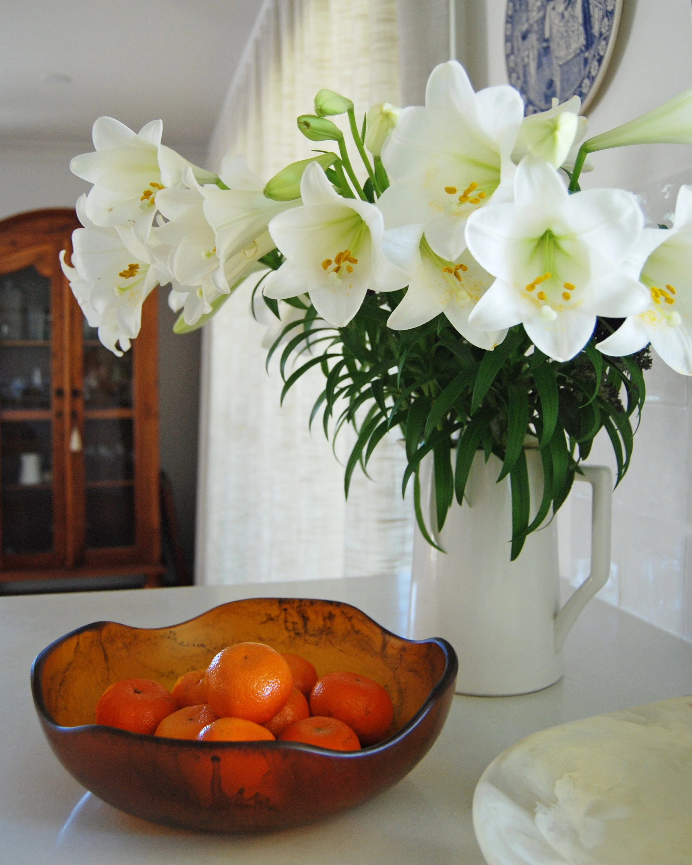 White lilies in a tall jug above a glass bowl of tangerines, beside a vintage timber cabinet — a quiet still life from the Oliv Interiors studio.