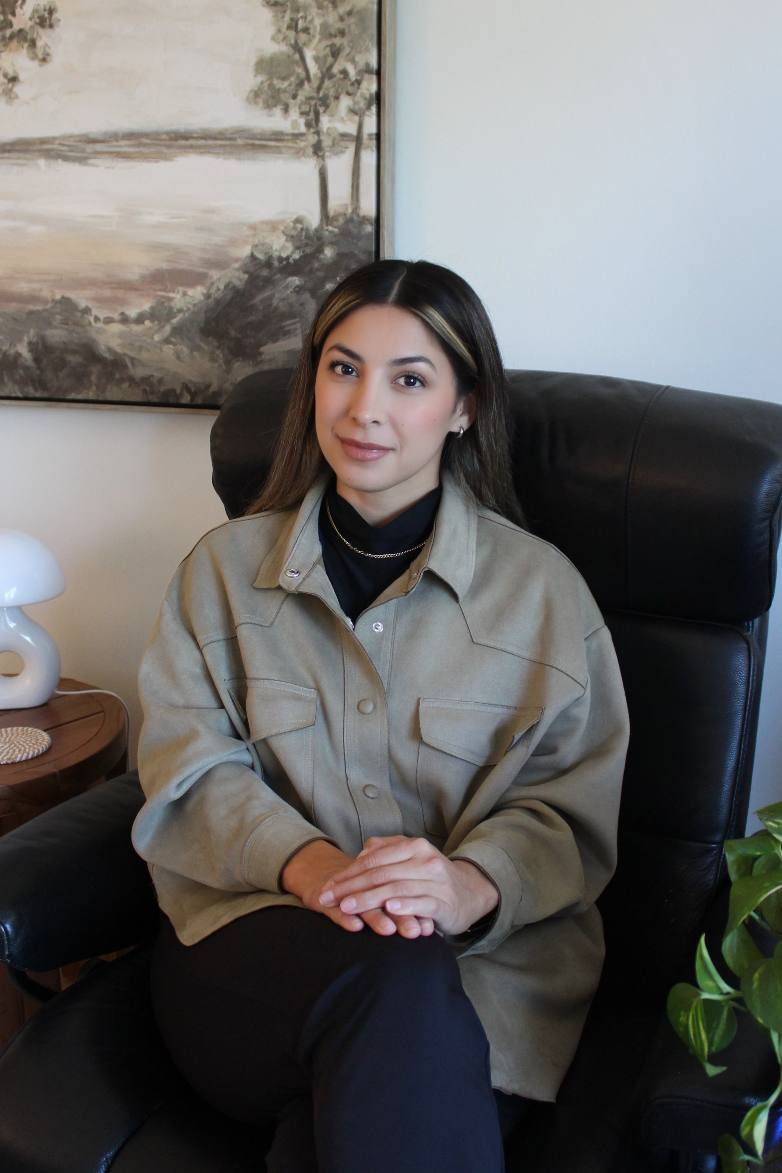A woman with long dark hair, wearing a beige jacket over a black shirt, sitting on a black leather chair in a room with a painting of trees and a landscape behind her.