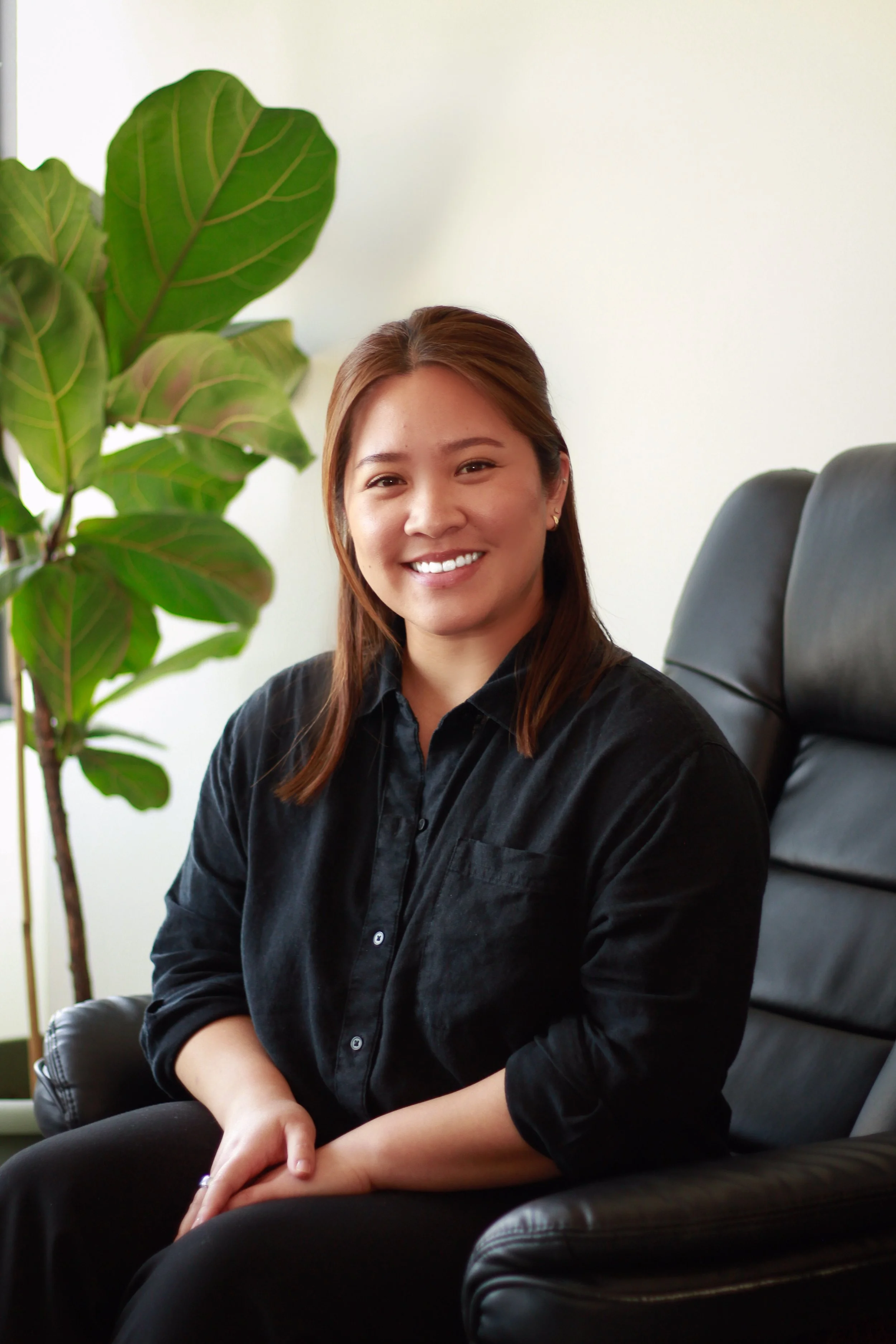 Woman sitting in a black leather chair, smiling, with a large green leafy plant beside her, in an indoor setting.