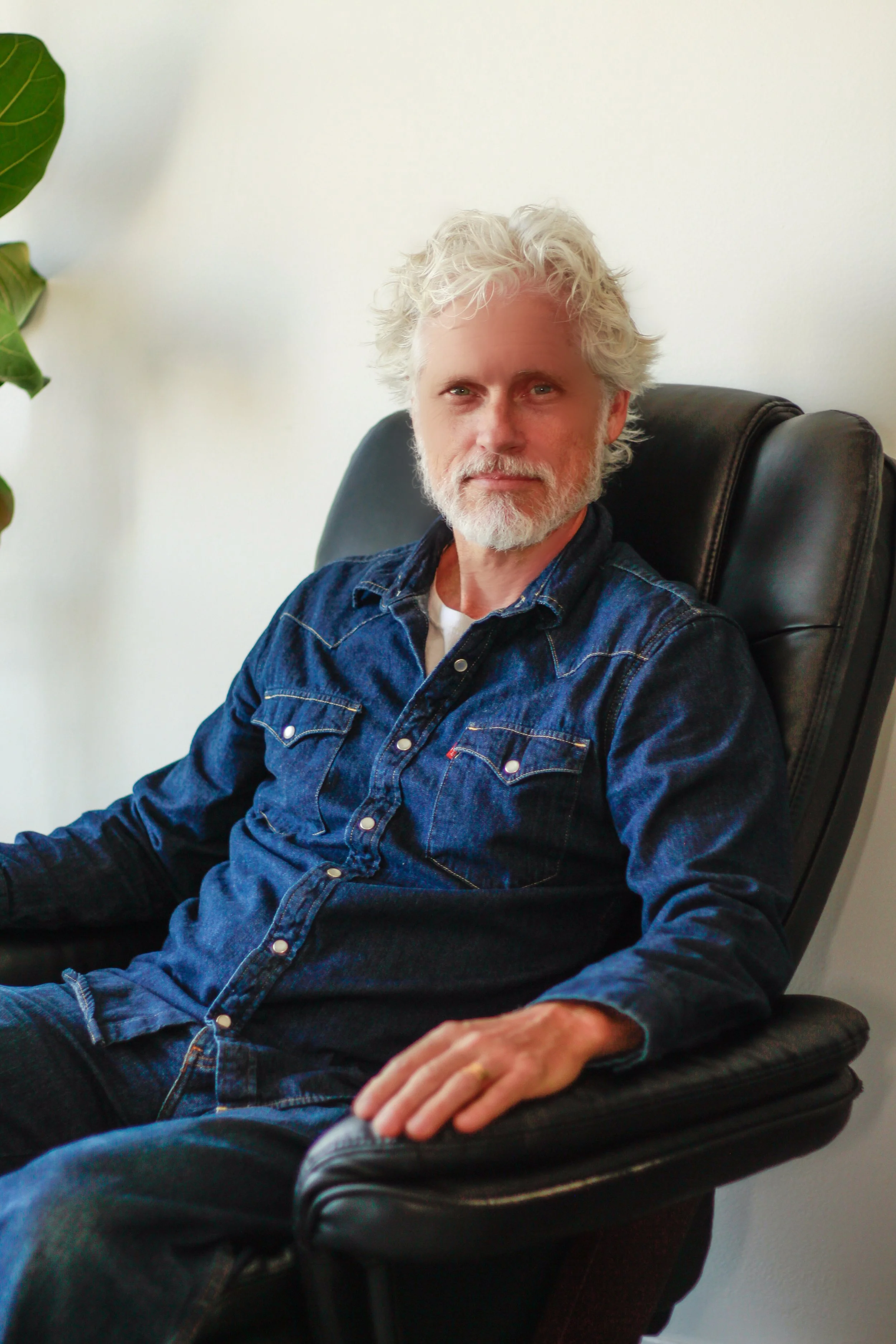 A man with white curly hair and a beard sitting in a black leather chair, wearing a dark denim shirt.