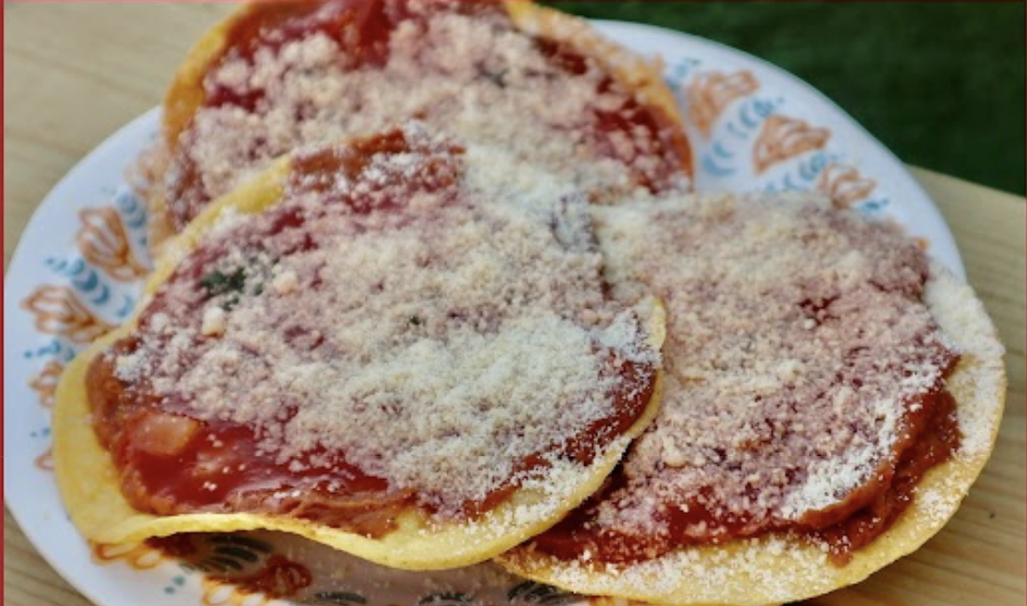 Three fried tortillas with a red sauce and shredded cheese on top of a paper plate sitting on a picnic table.