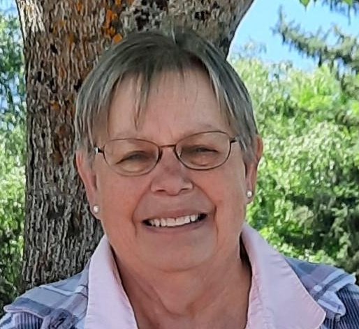 A smiling older woman with short gray hair and glasses, wearing earrings and a pink collared shirt under a light jacket, standing outdoors near a tree with green foliage in the background.