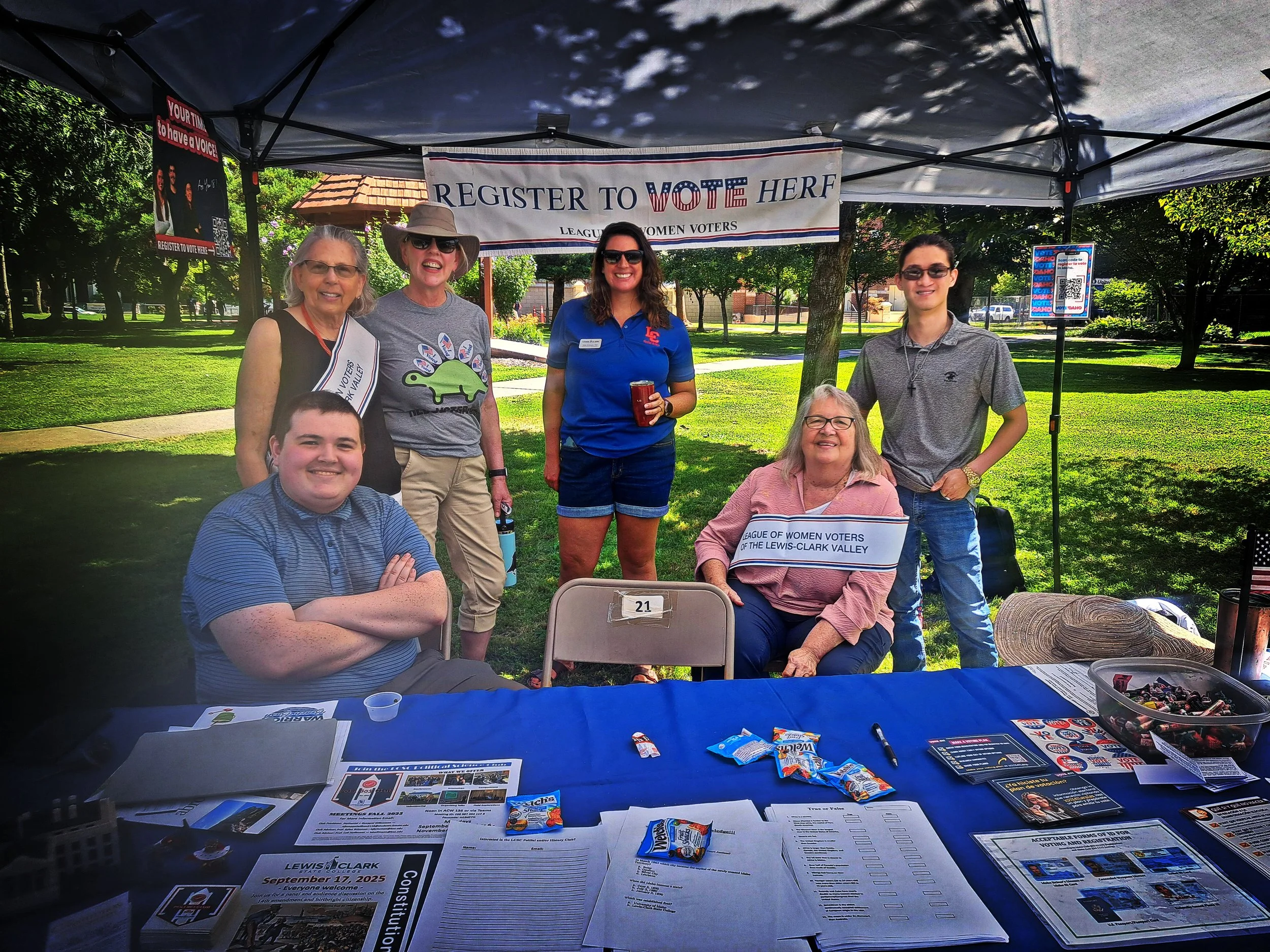 Group of people at a park registration booth for the League of Women Voters, with a banner that says "Register to Vote Here." There are four women and two men, some sitting and some standing, smiling at the camera. The table in front of them has informational papers, pamphlets, and snacks. Tents and trees are visible in the background.