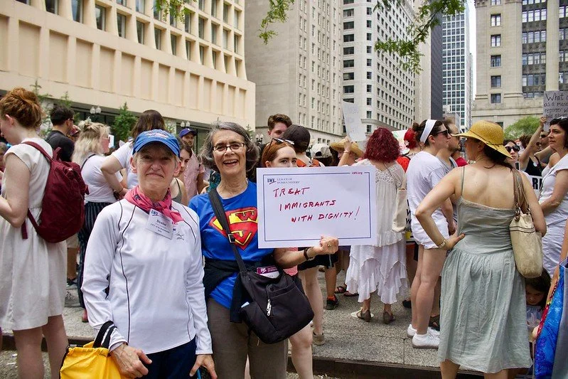 A diverse group of people gathered outdoors in a city, participating in a rally or protest, with one woman holding a sign supporting immigrant dignity.