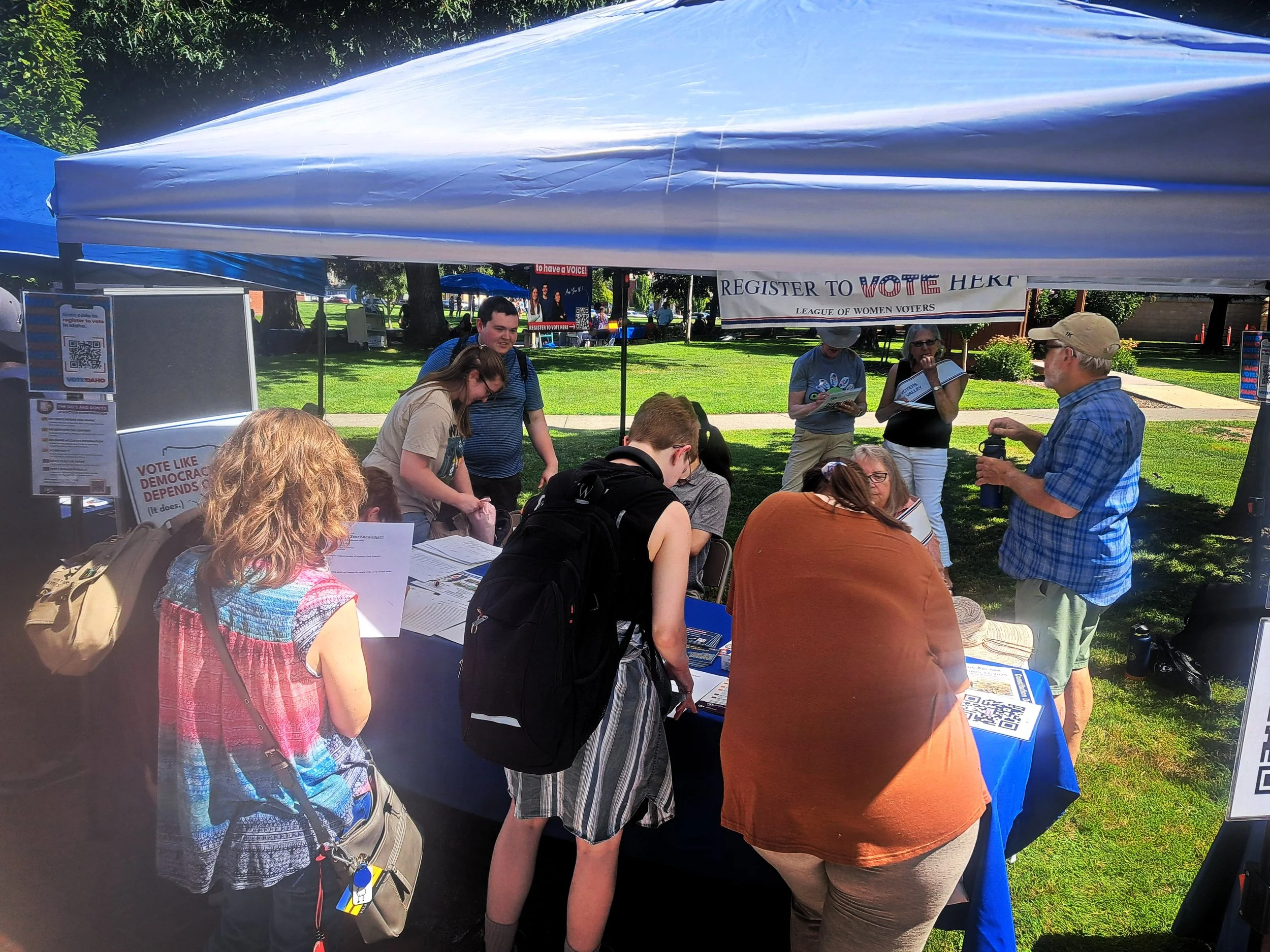 People gathered under a tent at a voting registration event in a park on a sunny day, with signs and tables set up for voter registration.