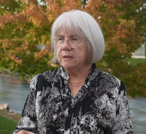 An elderly woman with white hair and glasses standing outdoors near a river with autumn-colored trees in the background.