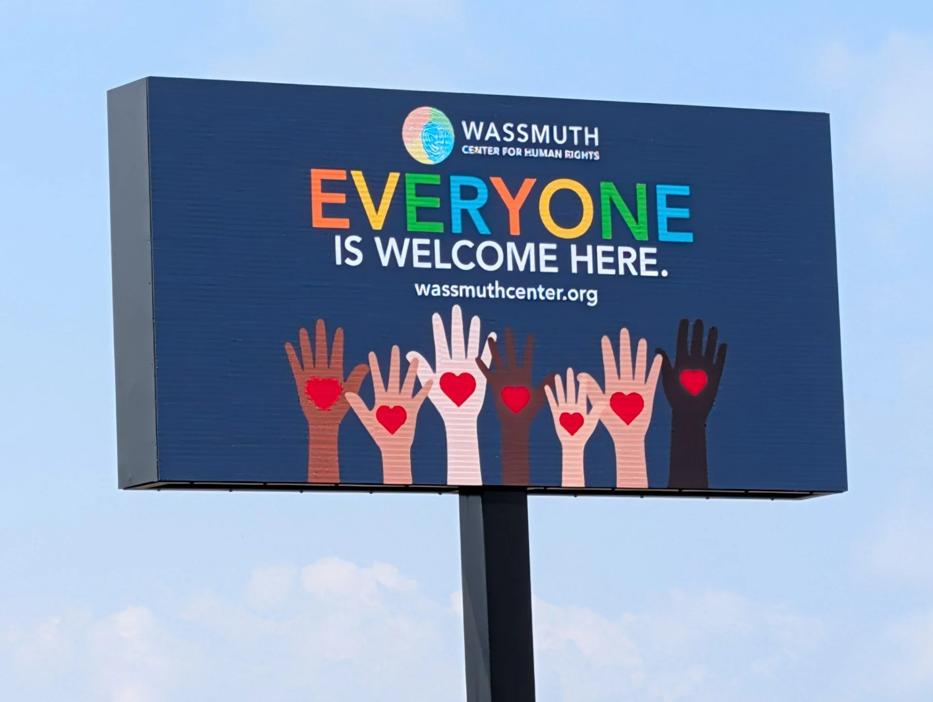 A billboard with the logo of Wassmuth Center for Human Rights, displaying colorful letters spelling 'Everyone is Welcome Here' and a row of six illustrated hands with red hearts on their palms.