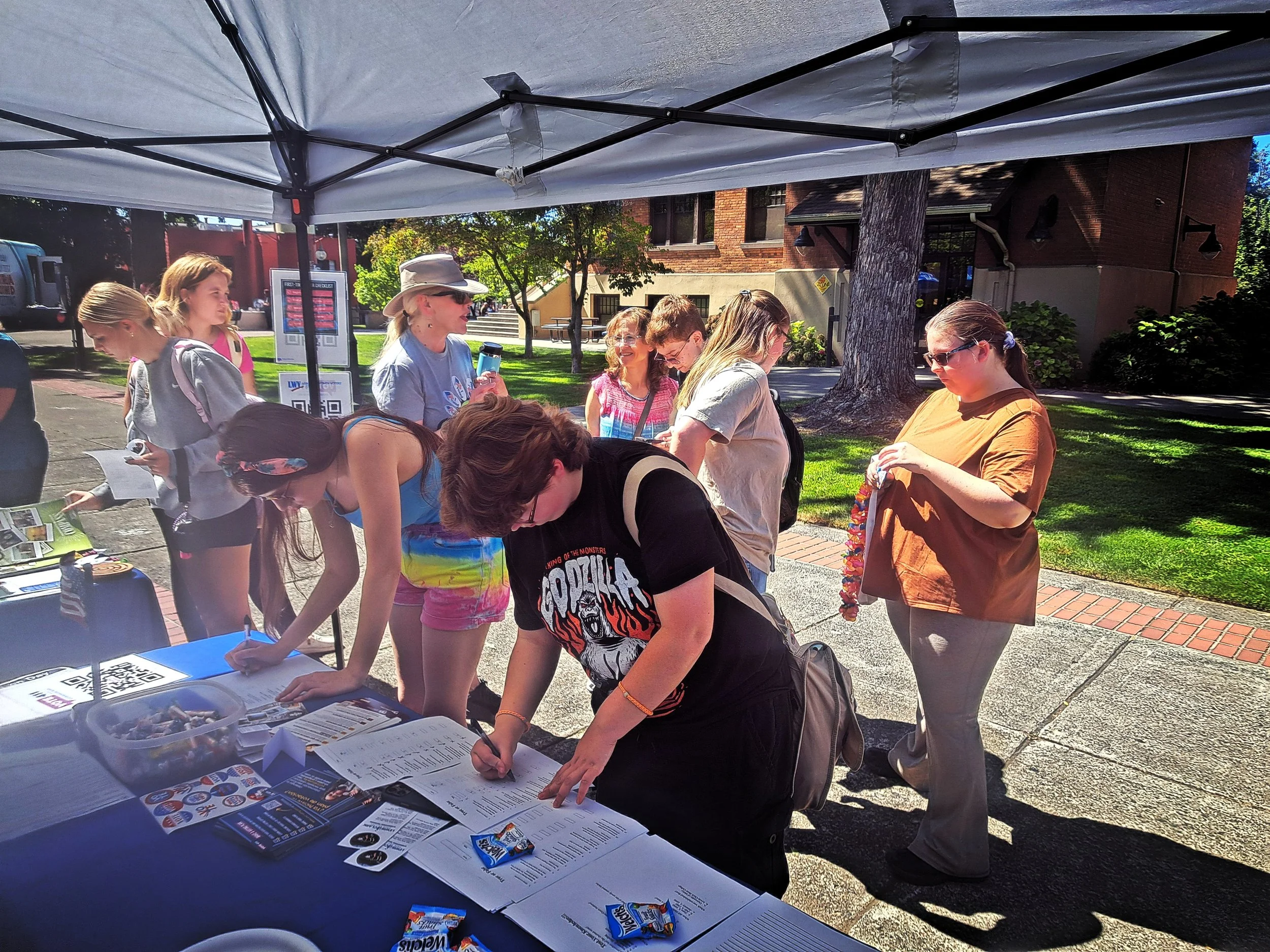 People lining up at an outdoor event registration booth under a canopy on a sunny day, with trees and a brick building in the background.