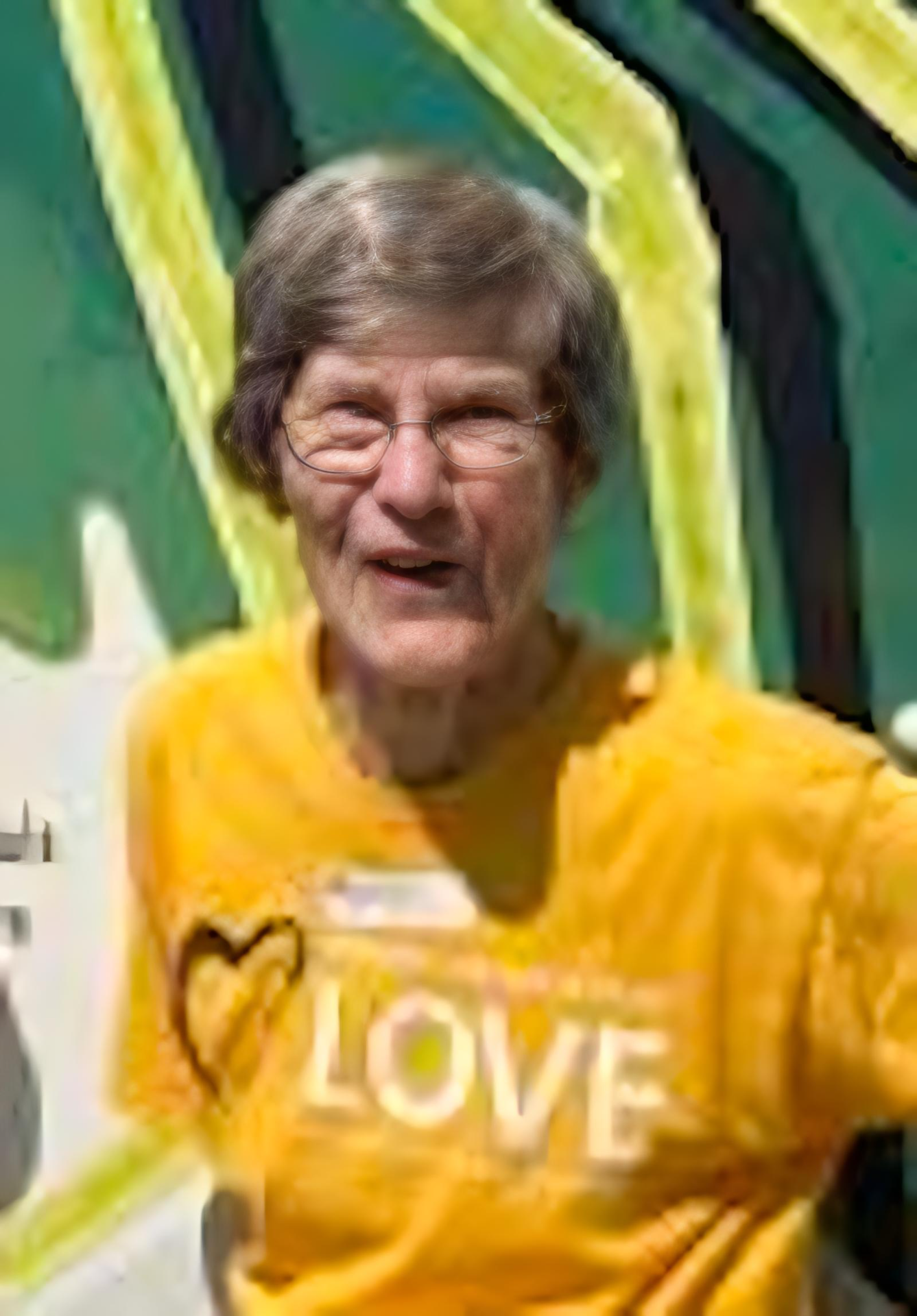 Older woman with glasses and gray hair smiling, wearing a yellow shirt with the word 'LOVE' and a heart on it, in front of a colorful painted background.