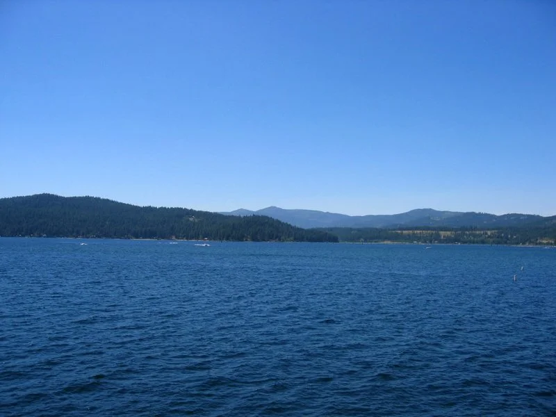 A large body of water with small boats, surrounded by forested hills and mountains under a clear blue sky.