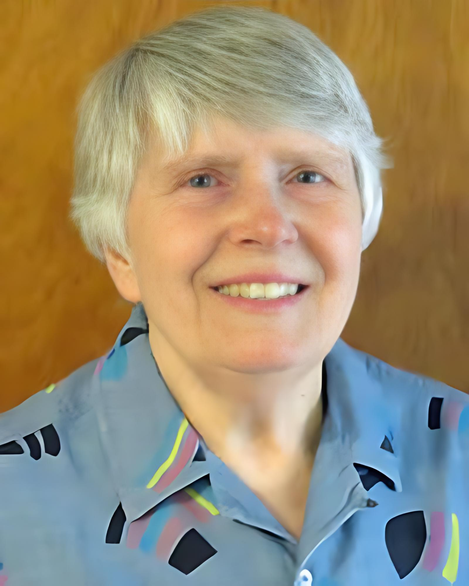 A woman with short gray hair wearing a gray shirt with a colorful geometric pattern, smiling, with a wooden background.