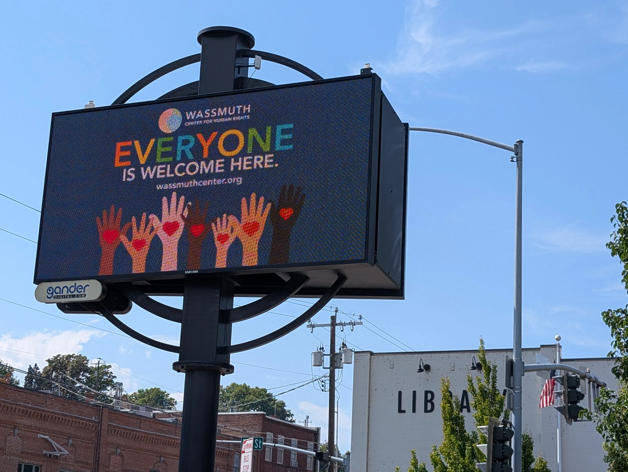 Digital billboard with a message promoting inclusivity and welcoming everyone, featuring colorful text and illustrations of diverse raised hands with hearts on palms.