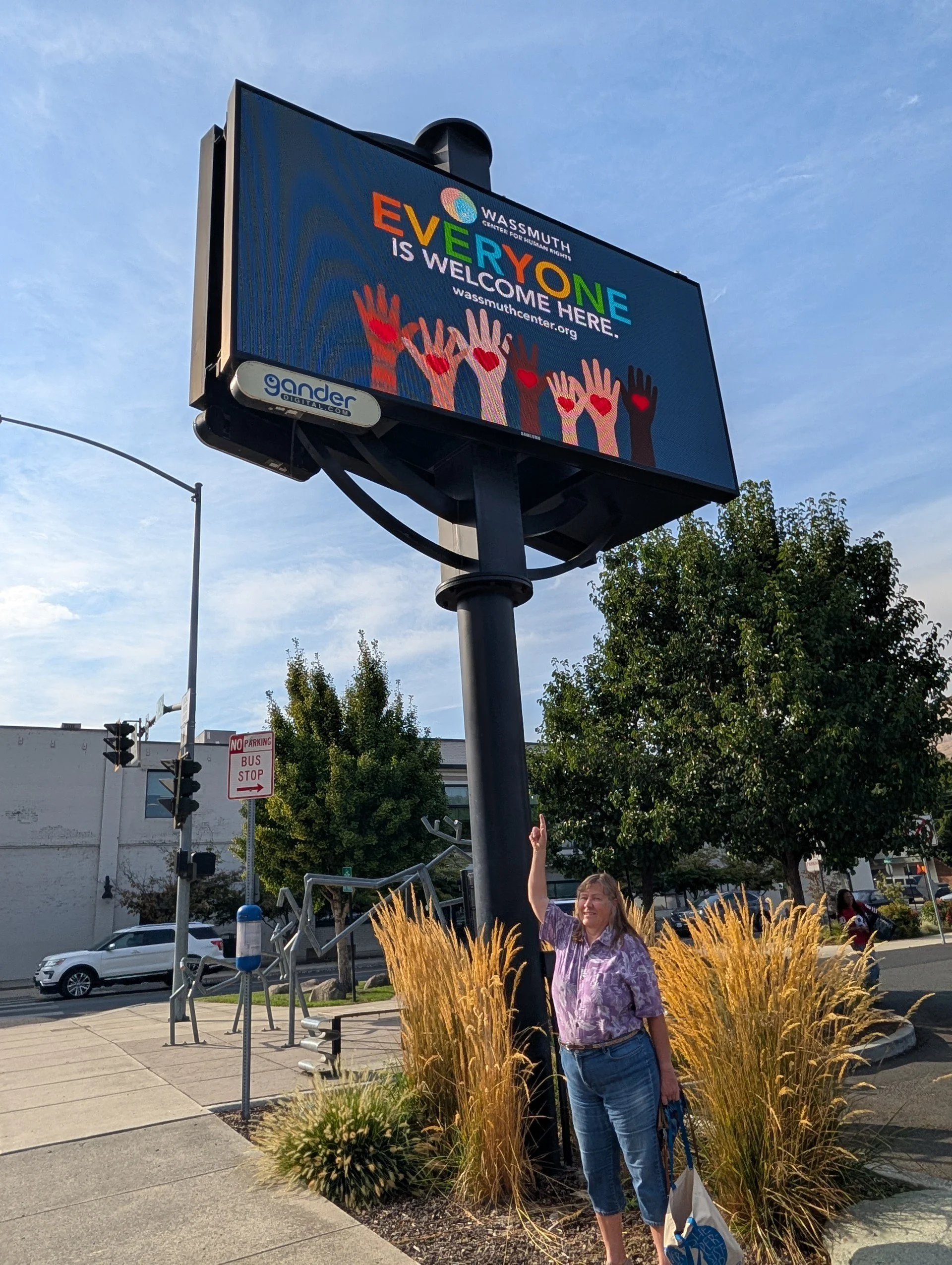 A woman standing next to a large digital billboard on a black pole, pointing upward. The billboard displays colorful hands with hearts on the fingertips, with text that reads 'Everyone is welcome here' for Was(s)mouth Center for Human Rights. The woman is wearing a purple floral blouse, blue jeans, and is holding a tote bag. The sidewalk has decorative grasses, trees, and street signs, with cars and buildings in the background.