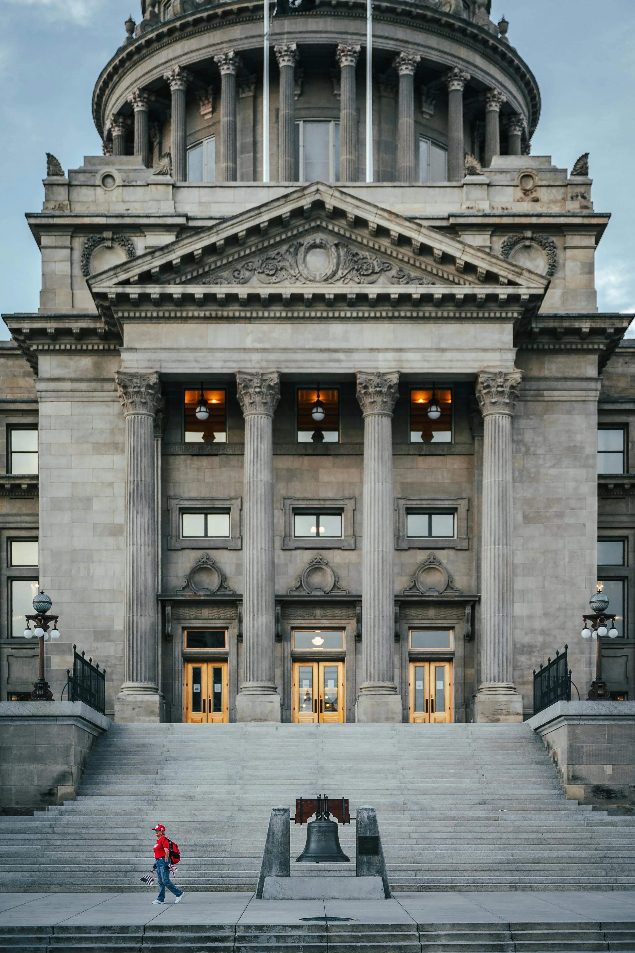 Front view of a historic government building with large columns, steps, and a bell in the foreground. A person wearing a red shirt and cap walks by.