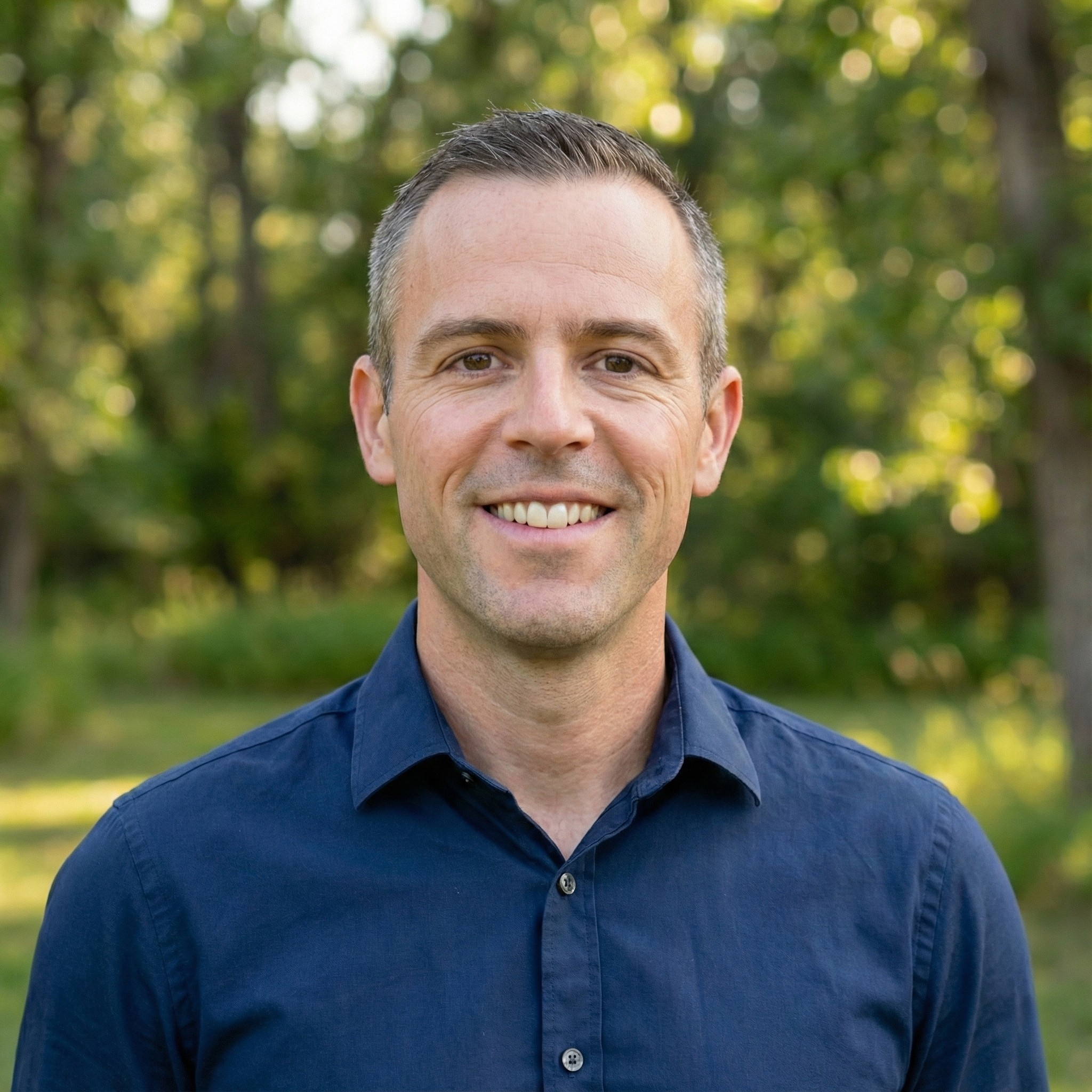 Smiling man in blue patterned shirt standing in front of trees.