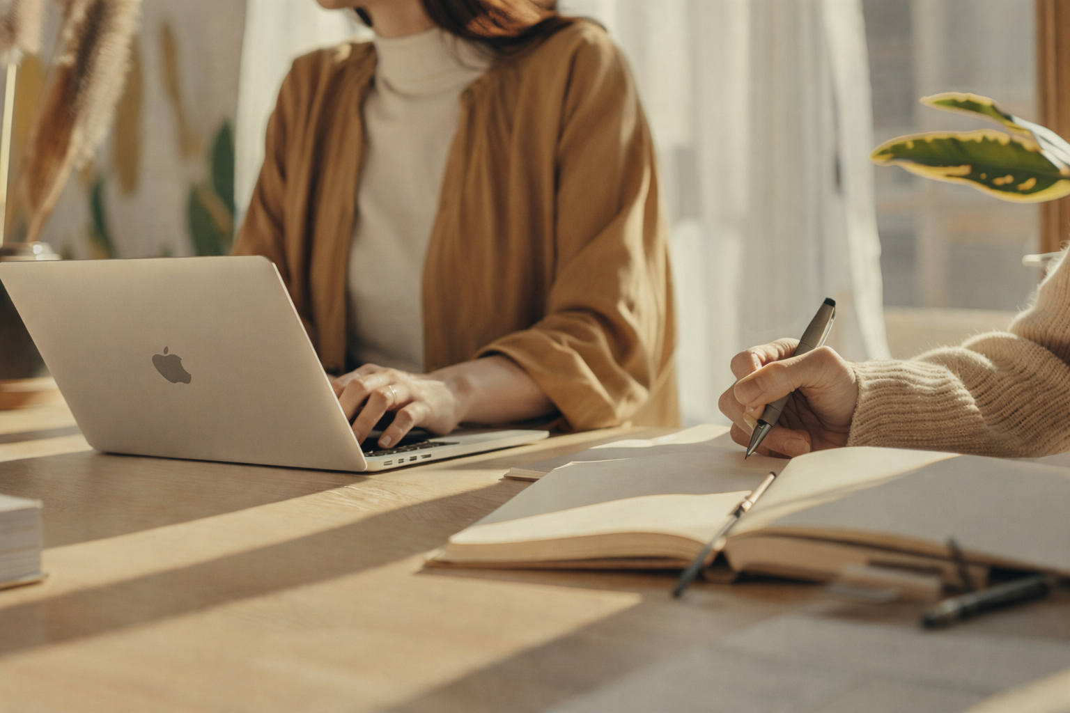 Two people working at a wooden table with a laptop and notebooks in a well-lit room with natural light.