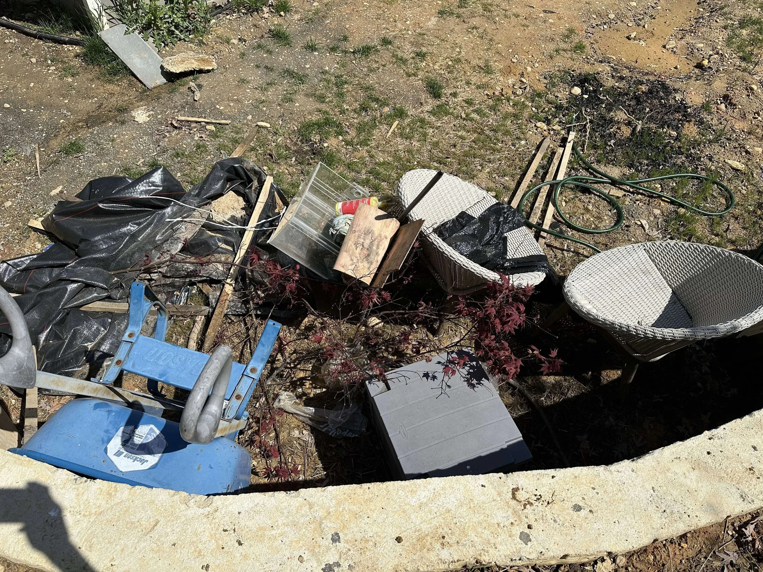 Various discarded patio furniture and gardening items on the ground, including two white wicker chairs, a child's blue ride-on toy, a gray plastic storage box, and miscellaneous debris.