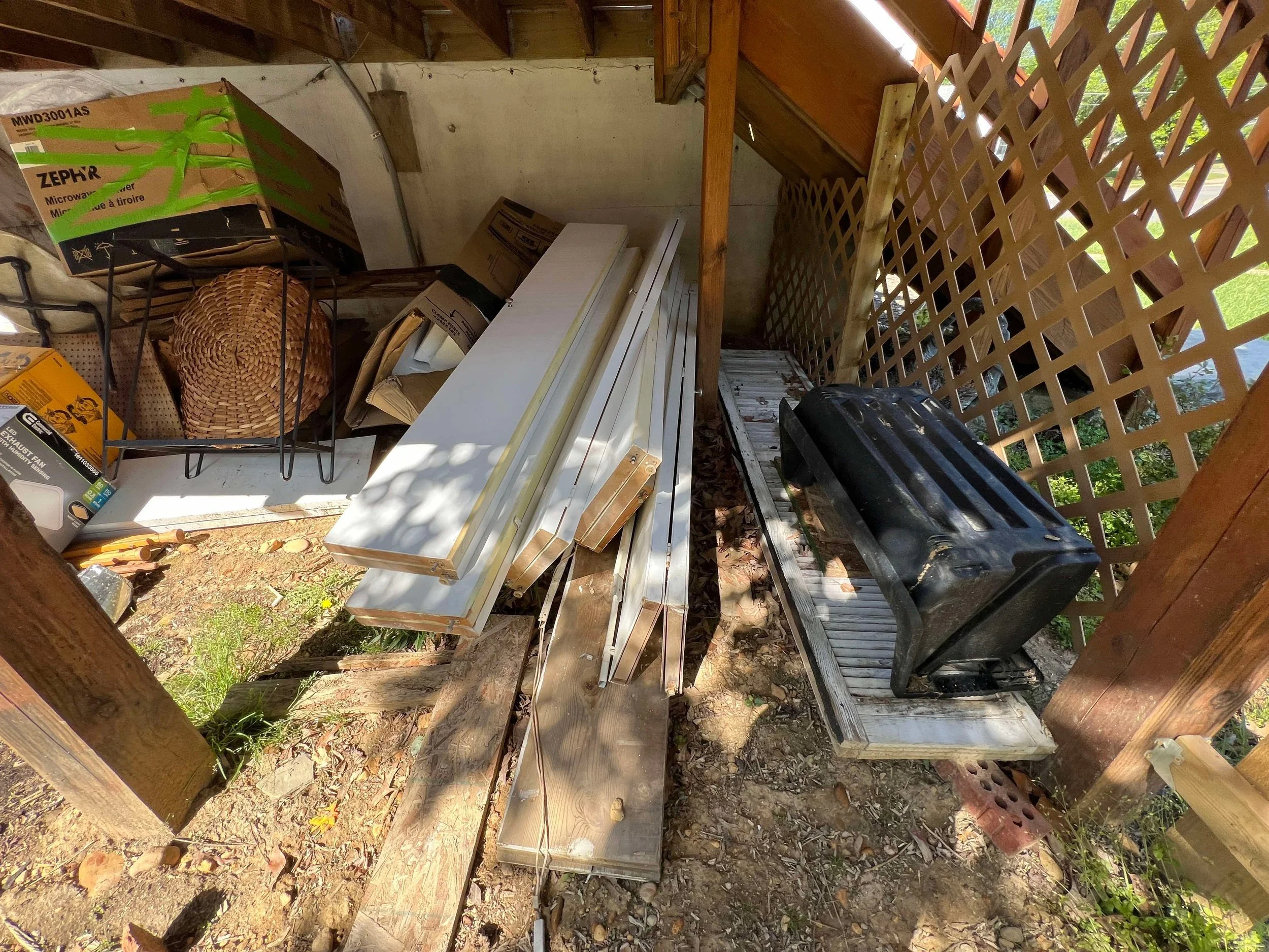 A small outdoor storage shed containing various household items, including a black plastic grill, white metal frames, long wooden boards, a wicker basket, a black wire basket with a box, and cardboard boxes, with lattice fencing on the side.