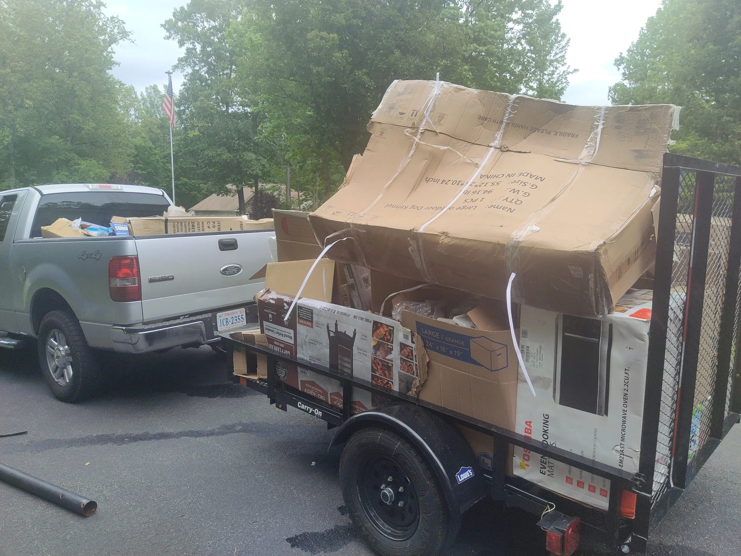 A pickup truck hitched to a trailer filled with large cardboard boxes, parked on a street with trees and an American flag in the background.