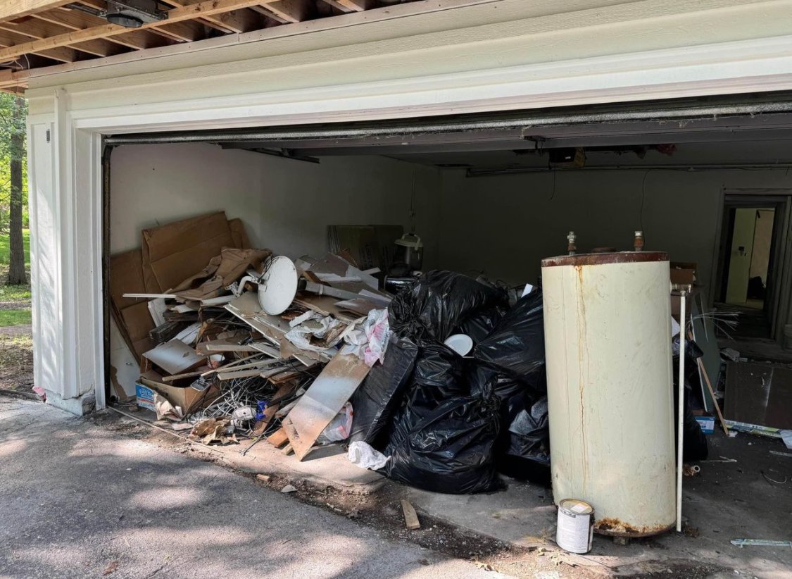 Garage filled with piled garbage, including cardboard boxes, black trash bags, an old rusty water heater, and miscellaneous household items.